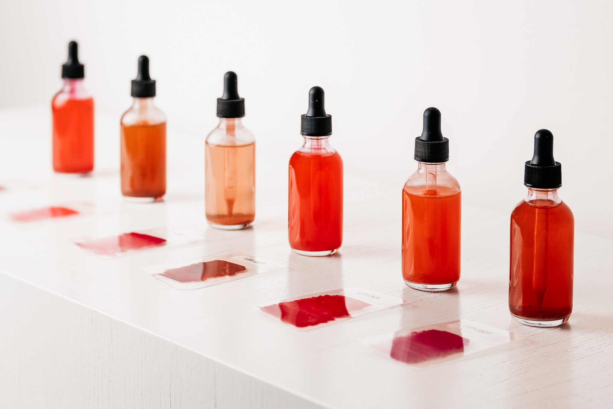 Row of glass dropper bottles with red liquid on a white surface, with color samples aligned beneath each bottle.