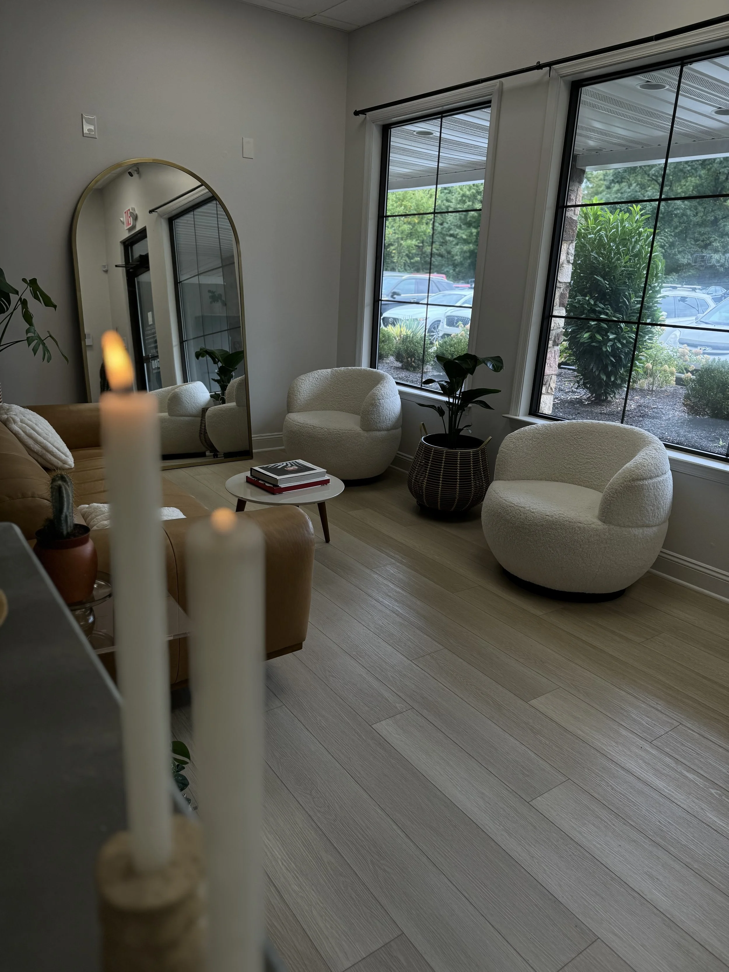Interior of a modern lounge with white curved chairs, a small round white table with books, large windows showing a parking lot and green trees outside, a tall plant in a basket, and a mirror reflecting part of the room. In the foreground, candles are blurred.