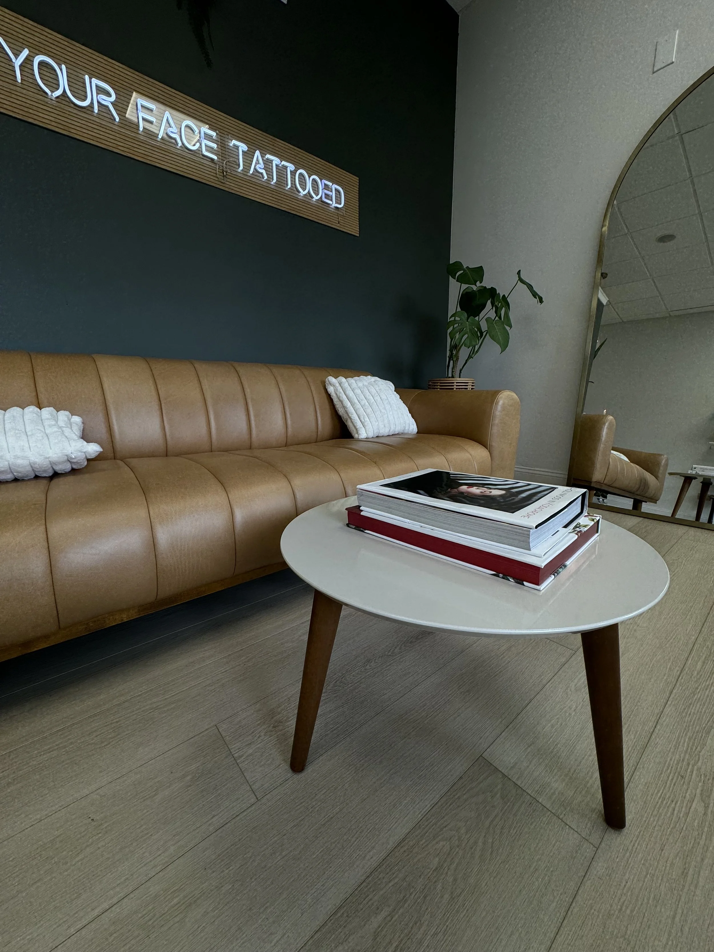 Modern waiting area with a tan leather sofa, white pillows, a small round table with magazines, a tall green plant, and a gray wall with a blue neon sign that reads 'YOUR FACE TATTOOED'.