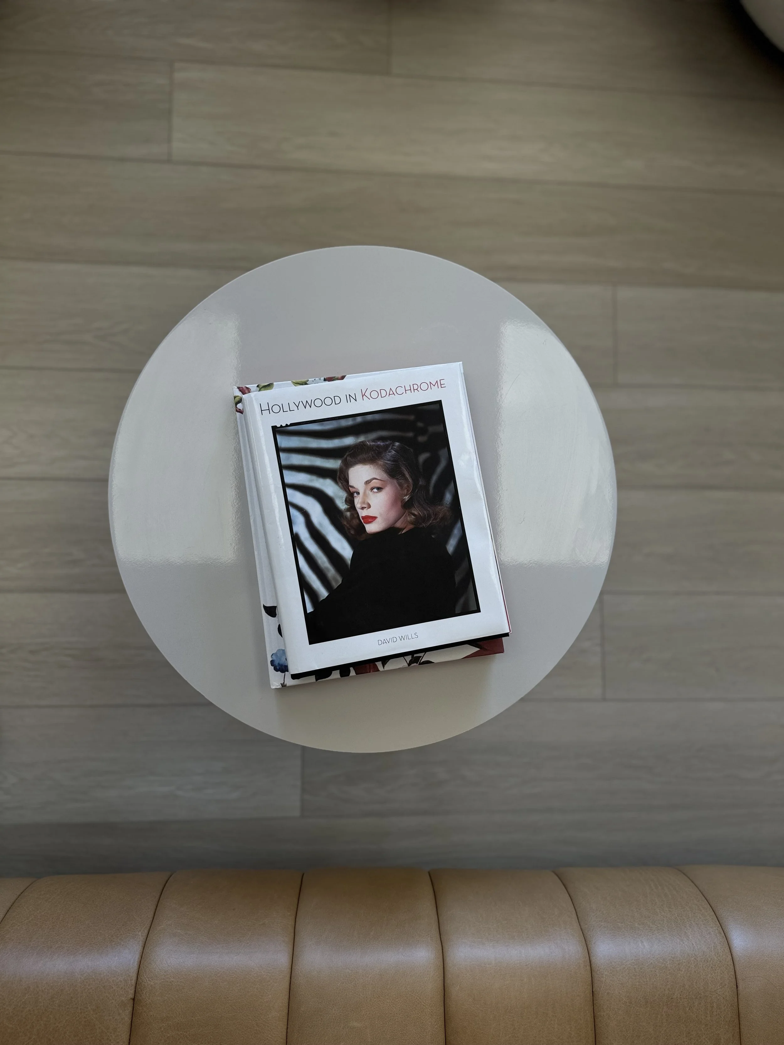 A white round table with a magazine on it, in a room with wooden flooring and a beige cushioned bench underneath.