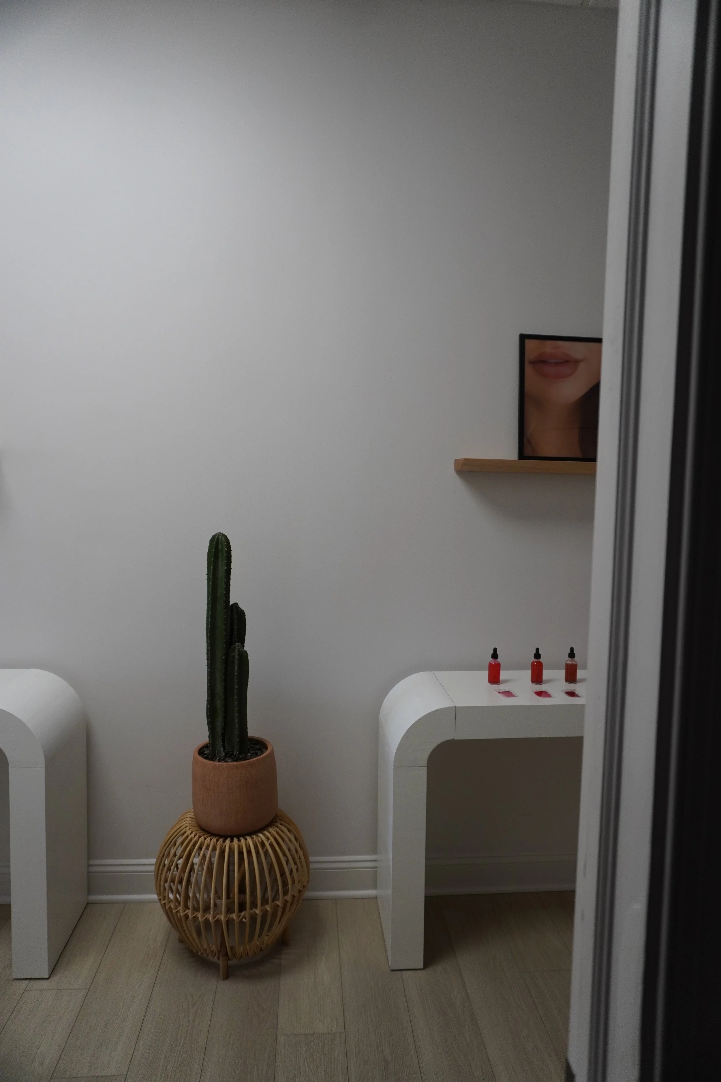 A white minimalist room with a tall cactus in a terracotta pot on a woven basket stand. There are two white modern side tables, each with three small bottles of nail polish in shades of pink and red. A framed picture of lips and part of a mirror are visible on the wall.
