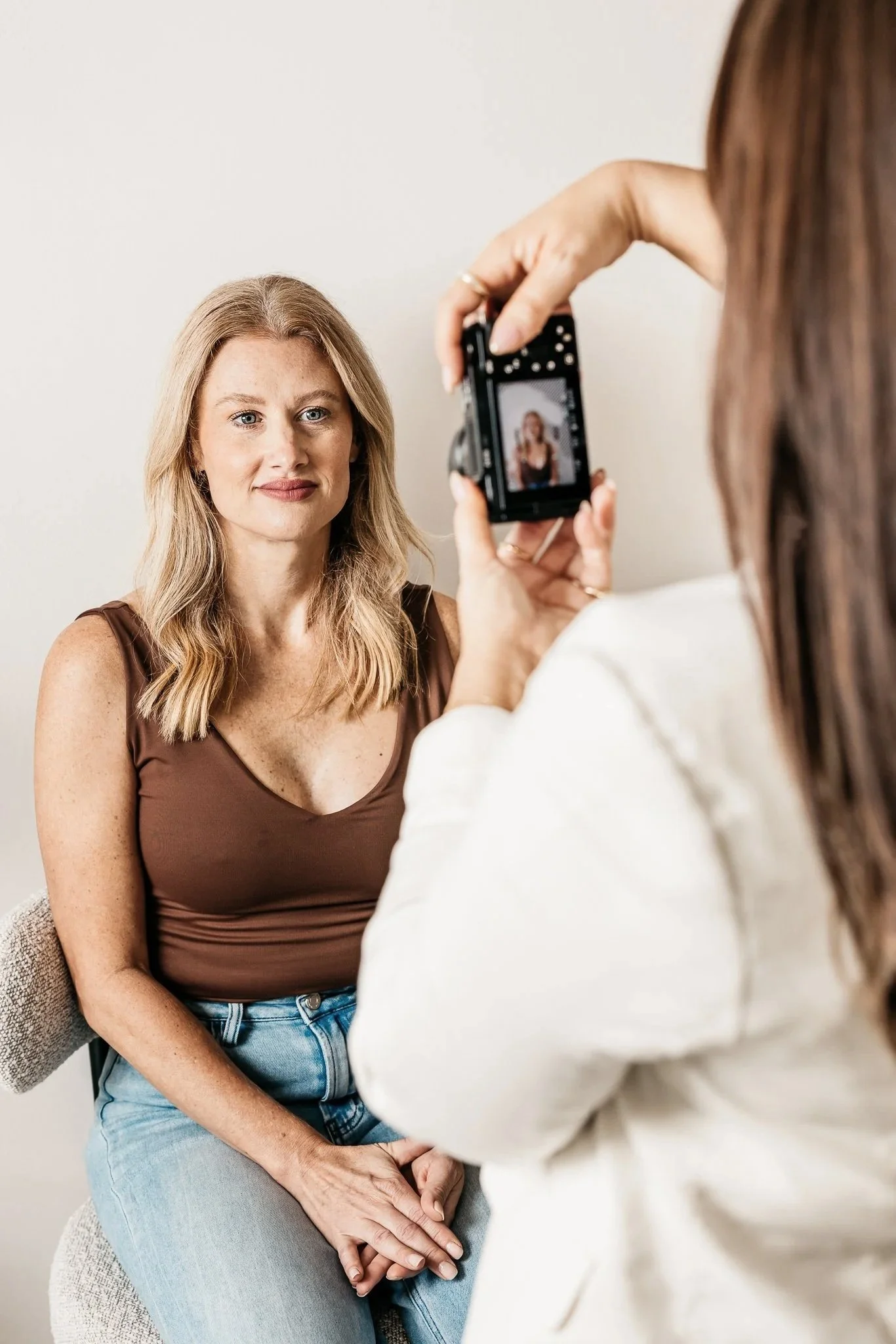 A woman with blonde hair and blue eyes sitting on a chair, being photographed by another woman with brown hair using a camera.
