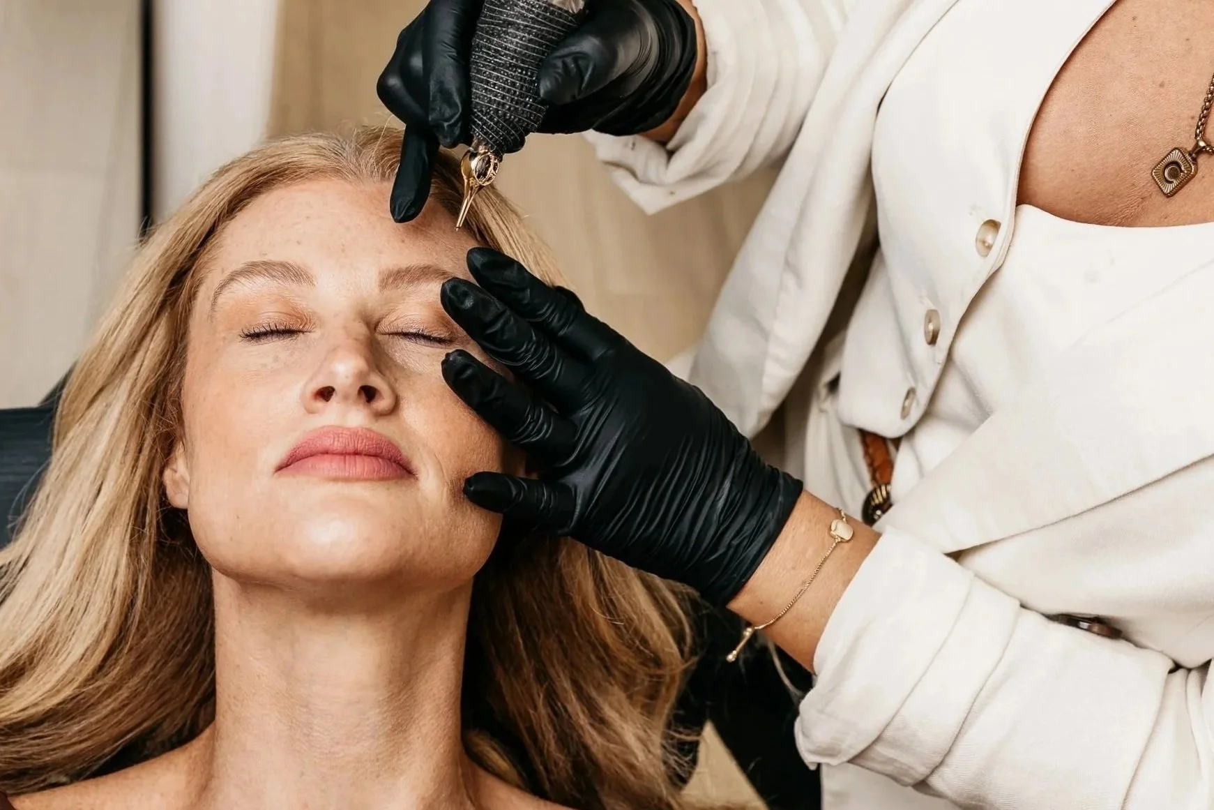 A woman with reddish hair and closed eyes receives a cosmetic or medical treatment on her face from a professional wearing black gloves. The professional is holding a small instrument near her forehead.