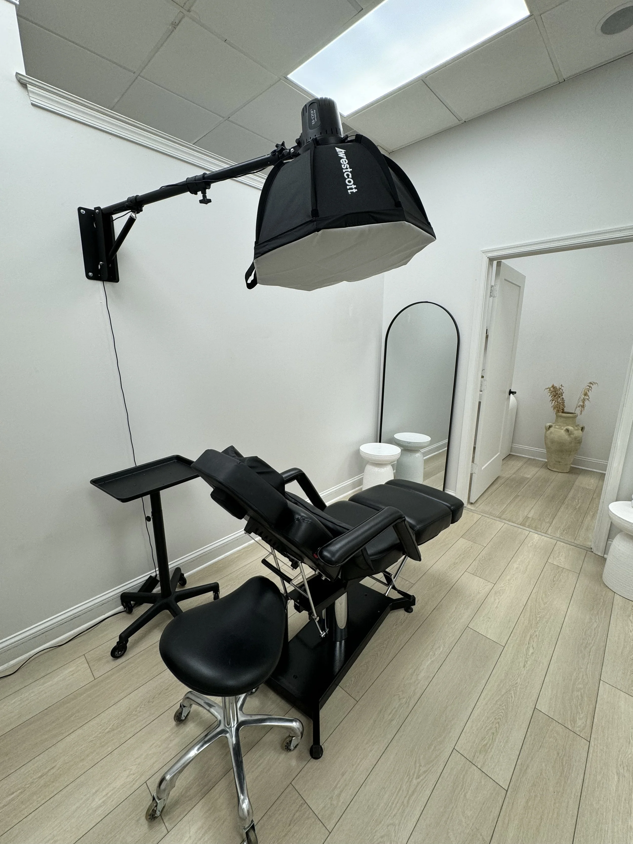 A medical examination room with a gynecological chair, a wall mirror, and a wall-mounted examination light. The room has neutral-colored walls, a light wood floor, and decorative vases in the corner.