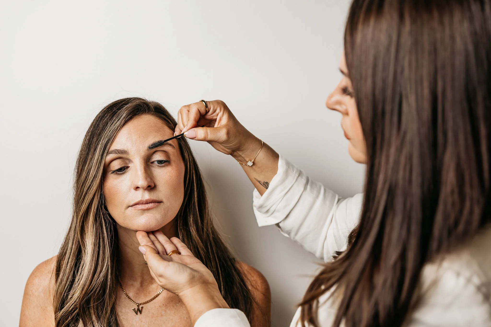 A woman with long brown hair getting her eyebrows shaped or waxed by a professional in a salon or spa setting.