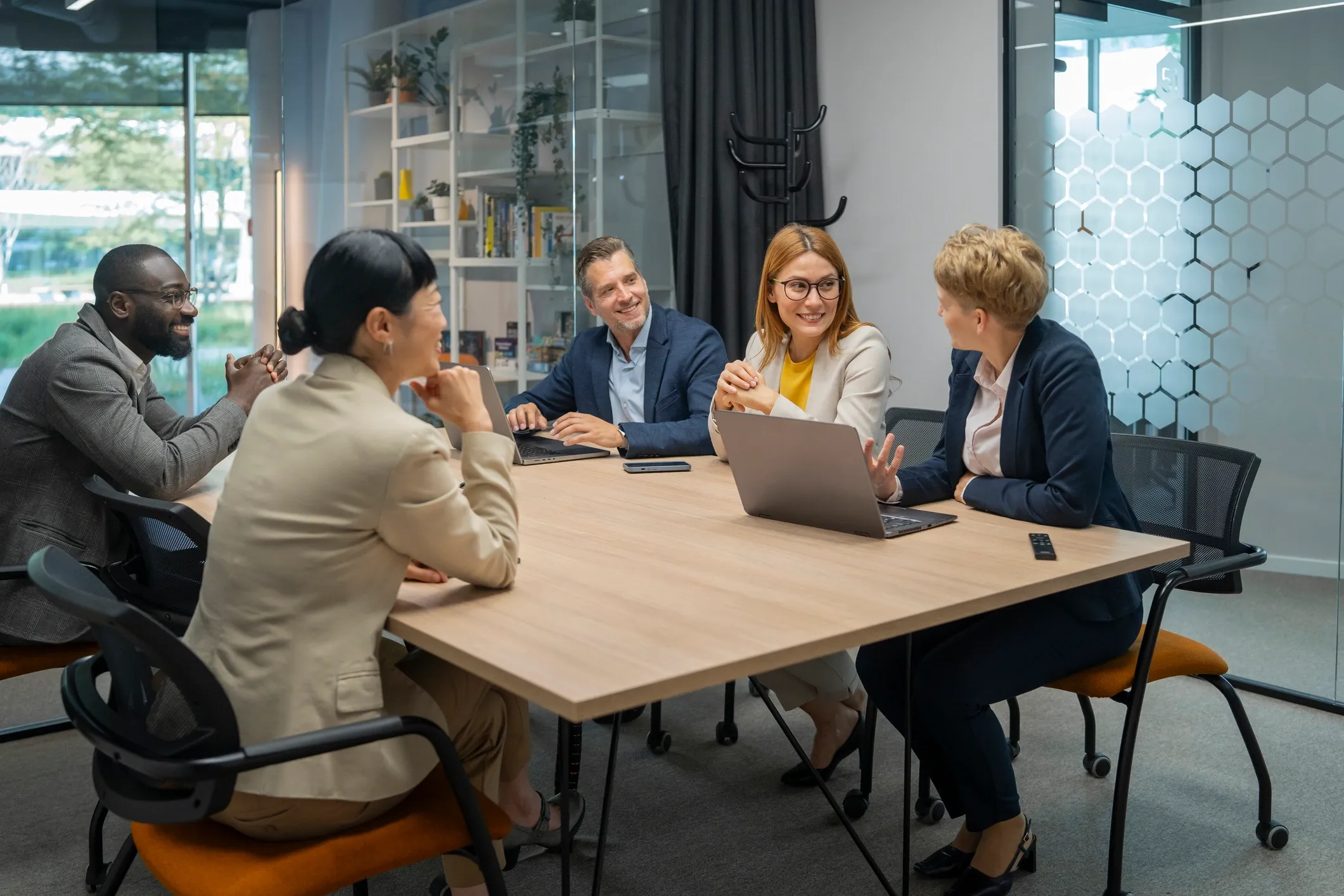 Six diverse professionals sitting around a conference table, smiling and engaged in a discussion, with laptops and a remote control on the table.