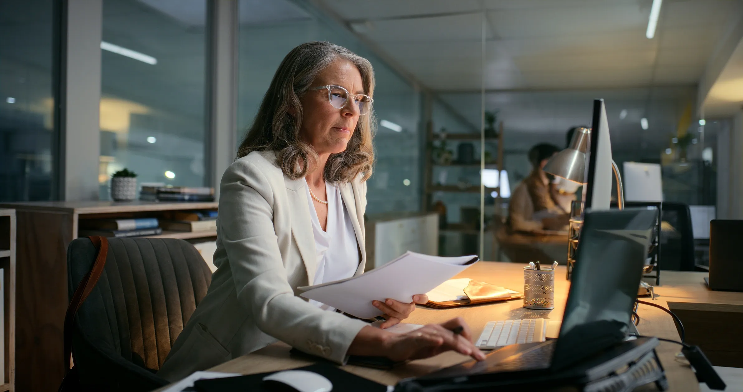 A woman with gray hair and glasses working at a desk in an office, looking at a computer screen and holding papers.