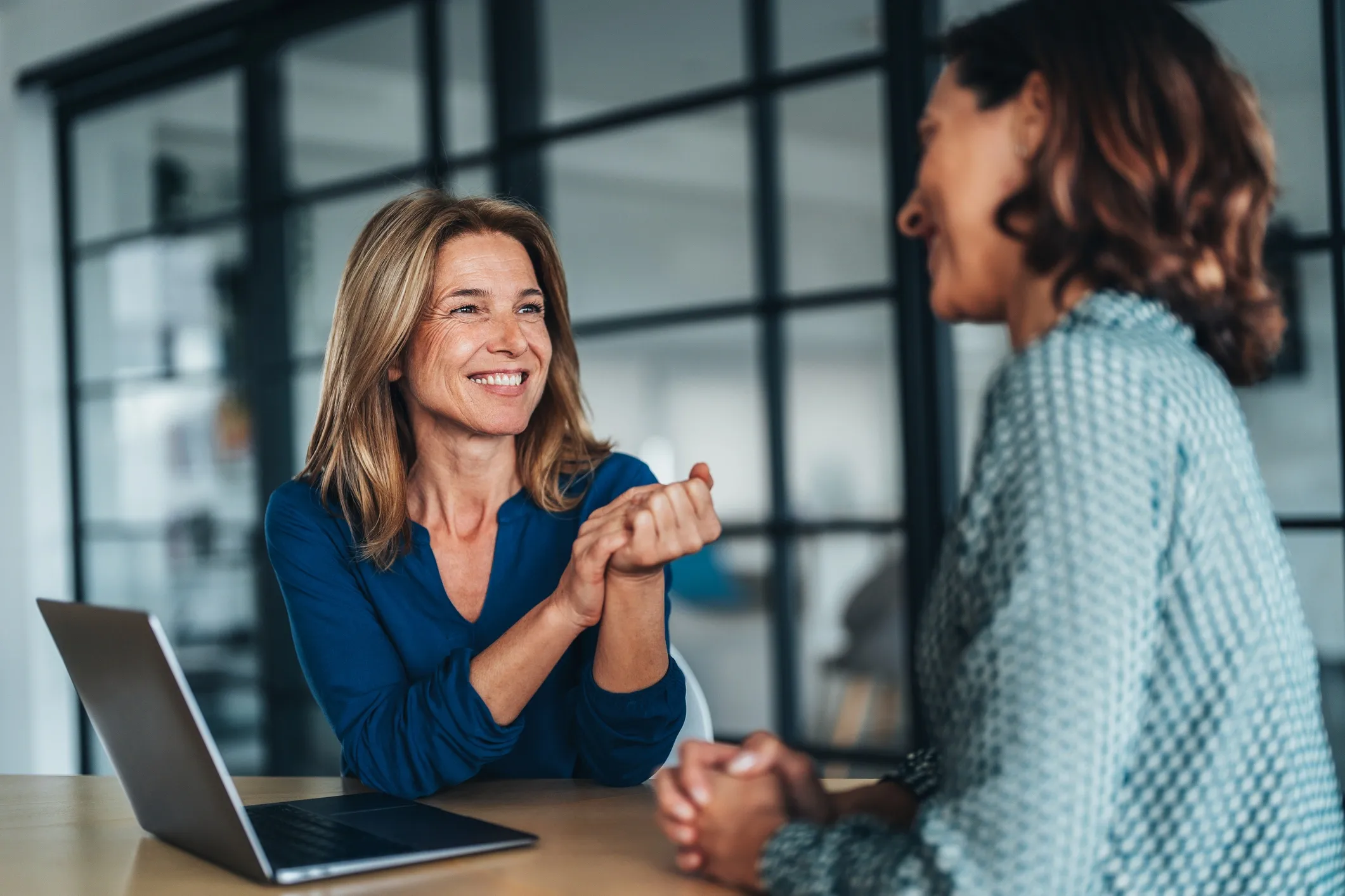 Two women sit at a table in a modern office, smiling and talking. One woman, with blonde hair, is wearing a blue blouse and has a laptop in front of her. The other woman, with dark hair, is wearing a patterned top.
