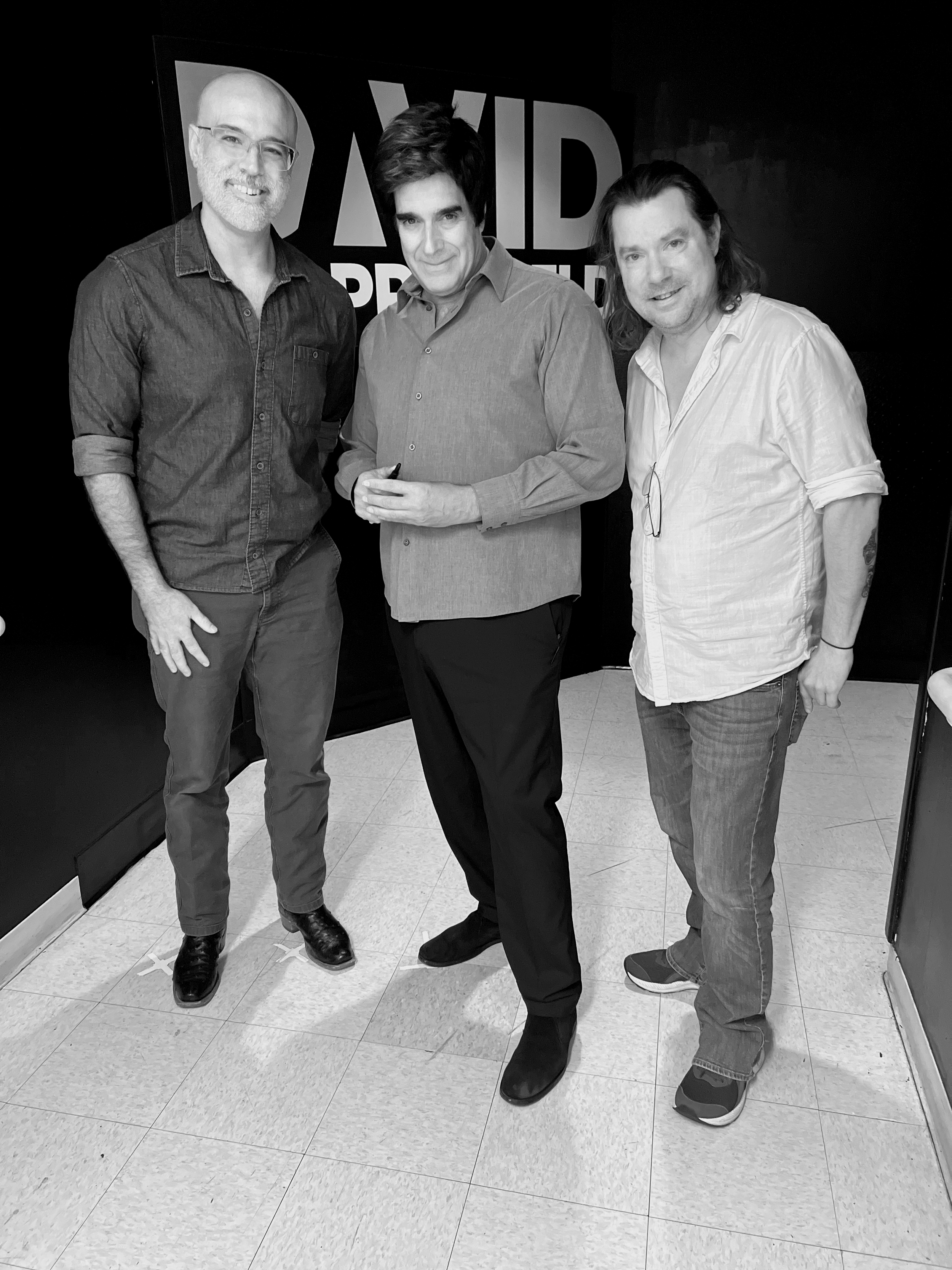 Three men standing together, smiling, in front of a black background with partial text "AID" visible. The men are J Weldon, Joshua Glantzberg, and David Copperfield.