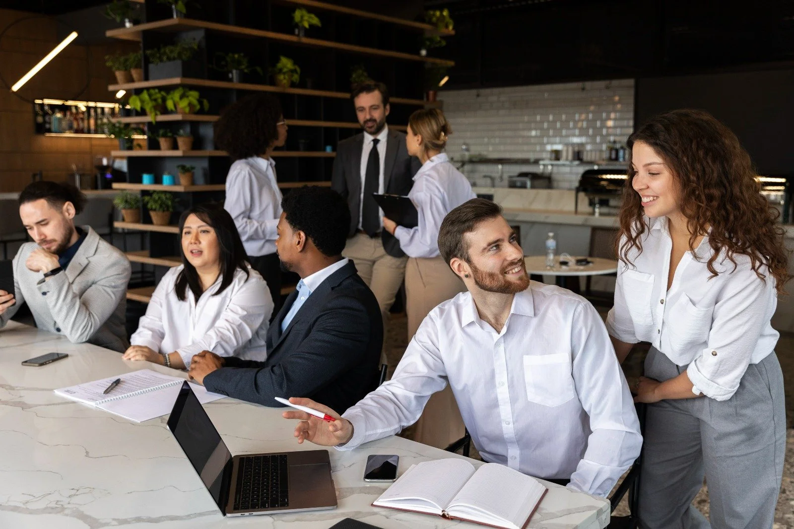 Business meeting in a modern cafe or co-working space with diverse people discussing and collaborating, some talking to each other at a marble table with laptops, notebooks, and smartphones, while others are in the background engaged in conversations.