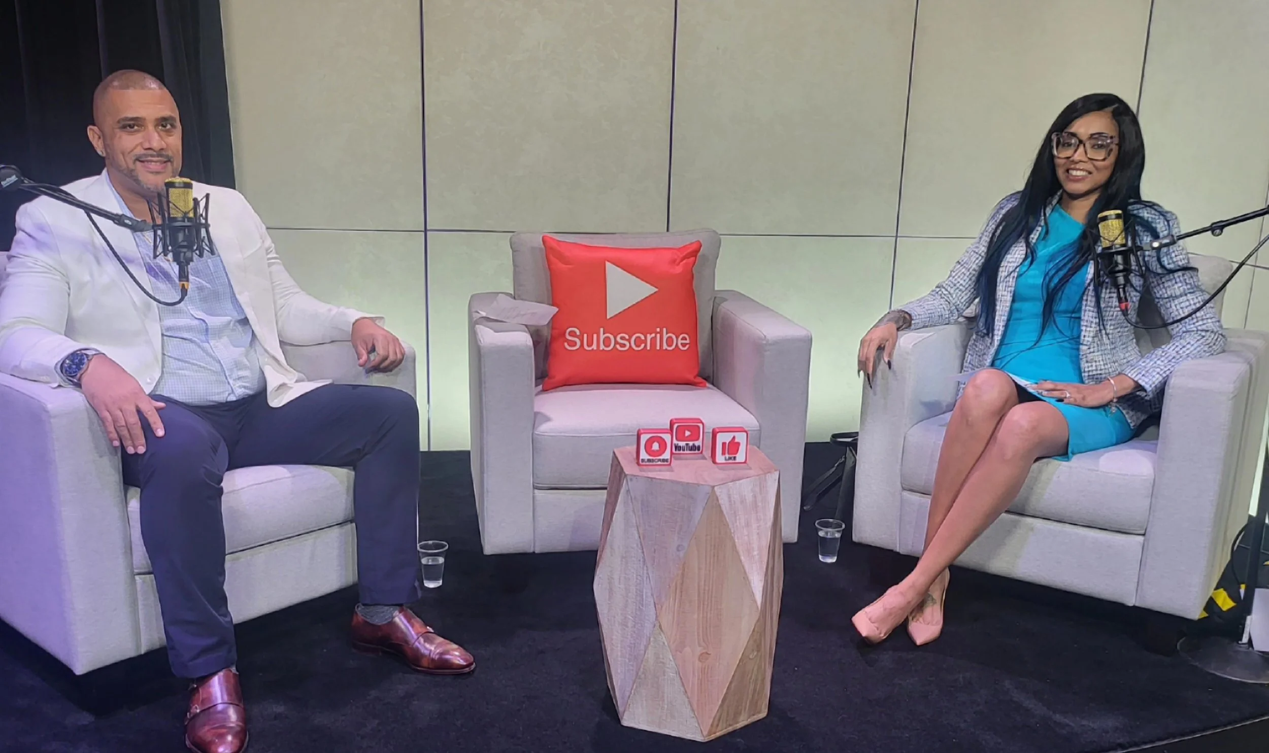 Two people sitting on armchairs in a podcast studio with microphones, a small wooden table with YouTube icons, and a red pillow with a YouTube play button and the word 'Subscribe' on it.