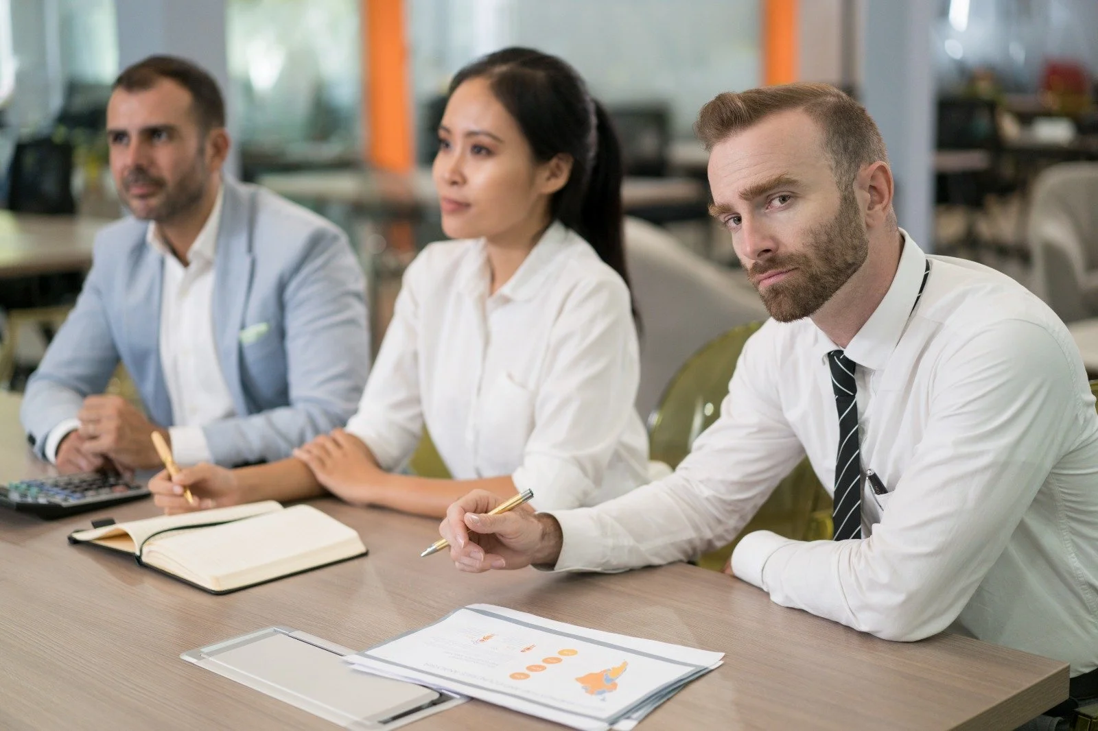 Three professionals sitting at a conference table with notebooks and documents, in a modern office setting.
