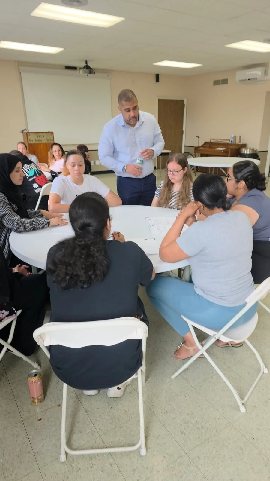 A group of women seated around a table engaged in a discussion, with a man standing and speaking to them in a room with a whiteboard and musical instruments in the background.