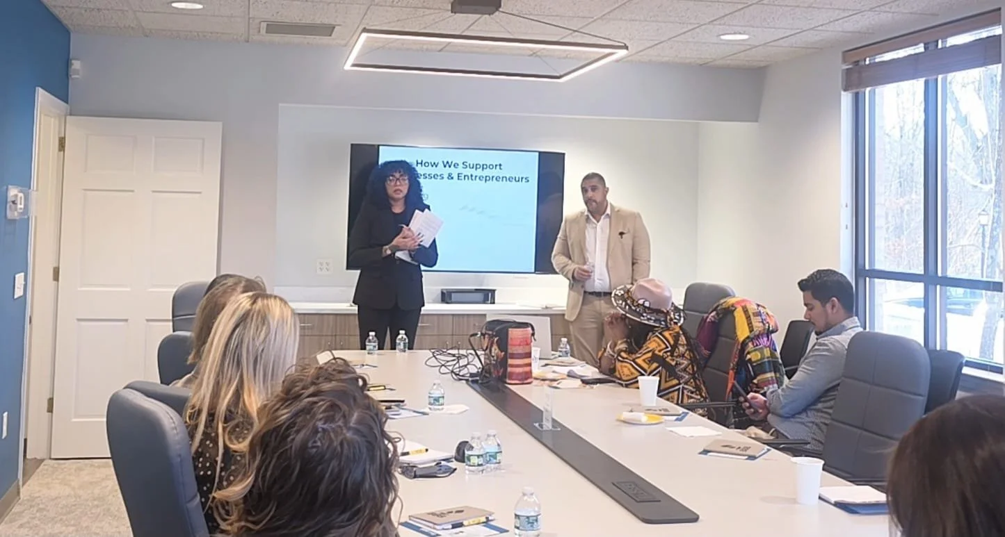 A business meeting in a conference room with people listening to a woman speaker and a man standing beside her. A screen behind them displays a presentation slide titled 'How We Support Businesses & Entrepreneurs.' Attendees are seated around a long table with water bottles, notebooks, and cups.