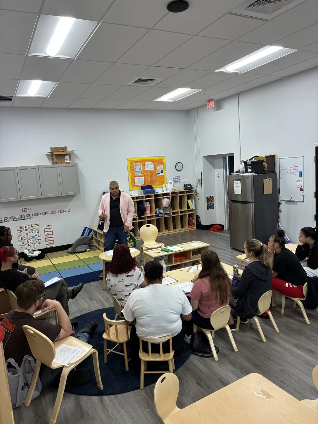 A classroom with a teacher standing in front of students seated around a semi-circle, white walls with orange bulletin board, gray cabinets, refrigerator, and whiteboard.