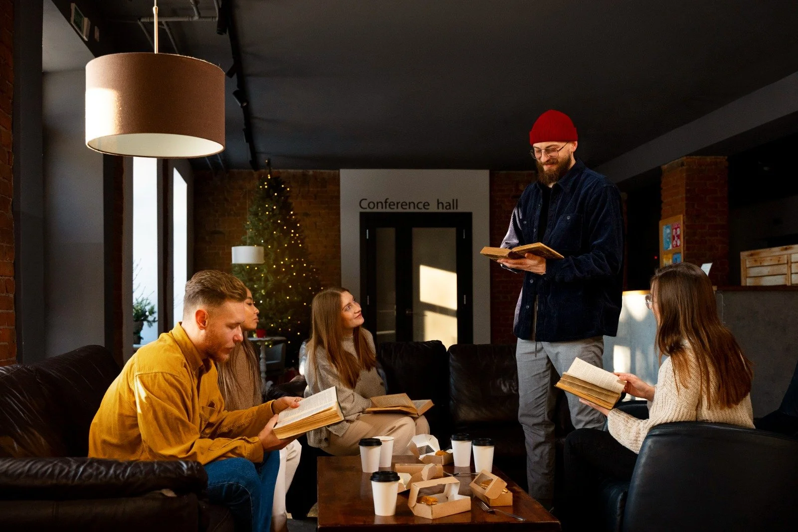 A group of five young adults sitting on a black leather couch in a modern conference room with exposed brick walls and large windows, while a man in a red beanie, glasses, and dark jacket stands reading from a book. The room is decorated with a Christmas tree with lights, coffee cups, and food boxes on a wooden table.