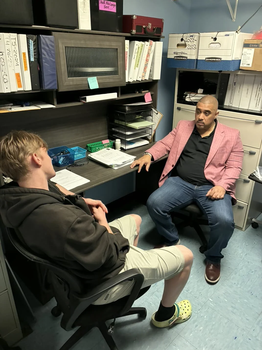 Two men having a conversation in an office. One is sitting at a desk and the other is sitting in a chair across from him. The office has shelves with binders, boxes, and office supplies.