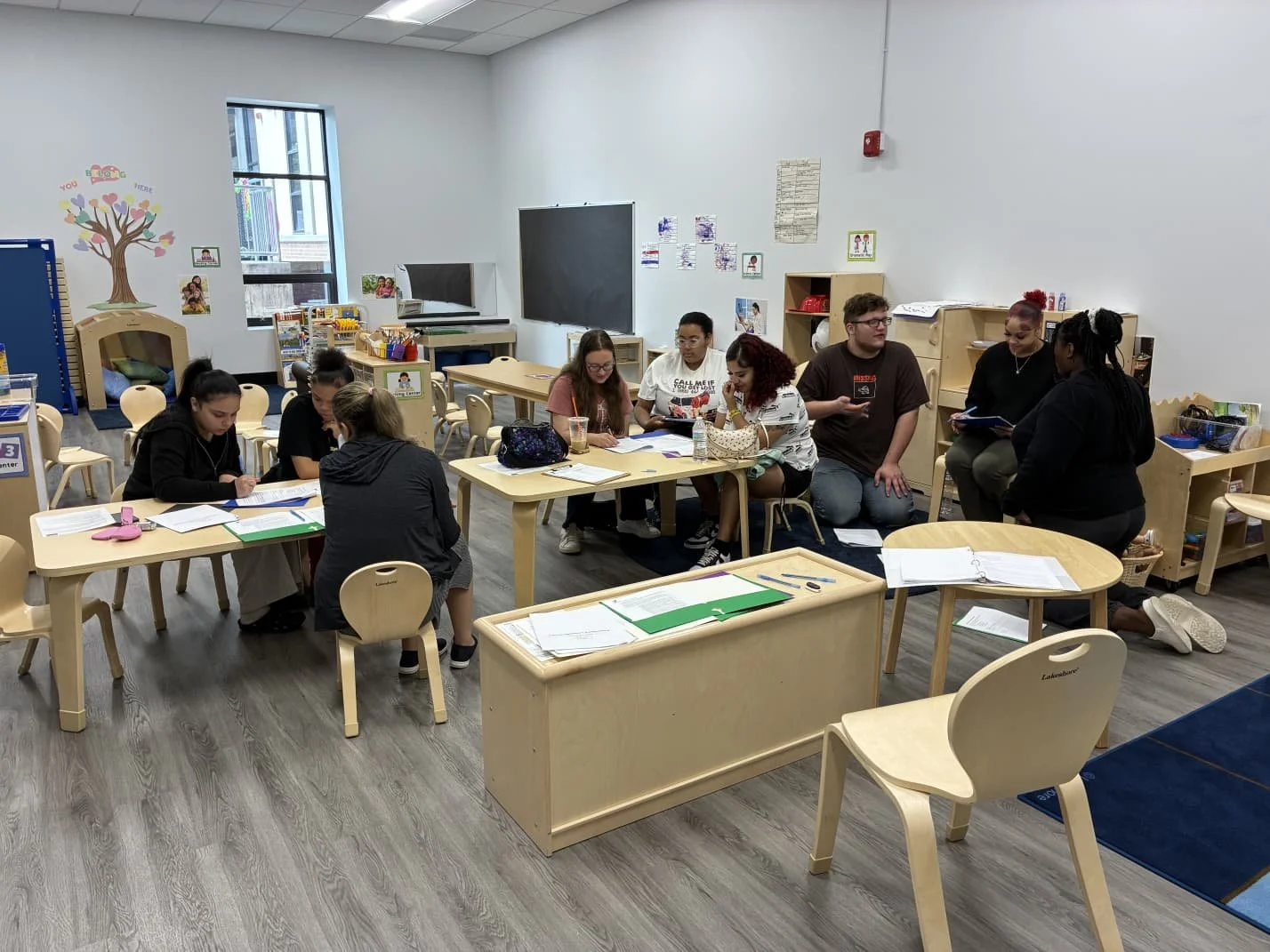 A group of people, likely students and a teacher, gathered in a classroom with educational posters, a blackboard, and small chairs and tables, some with papers and supplies on them.