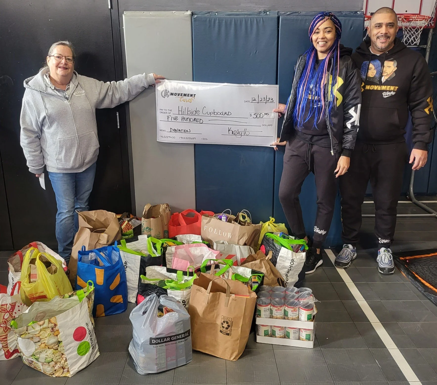 People holding a large donation check for $500 in front of grocery bags and canned goods, with a basketball hoop in the background.