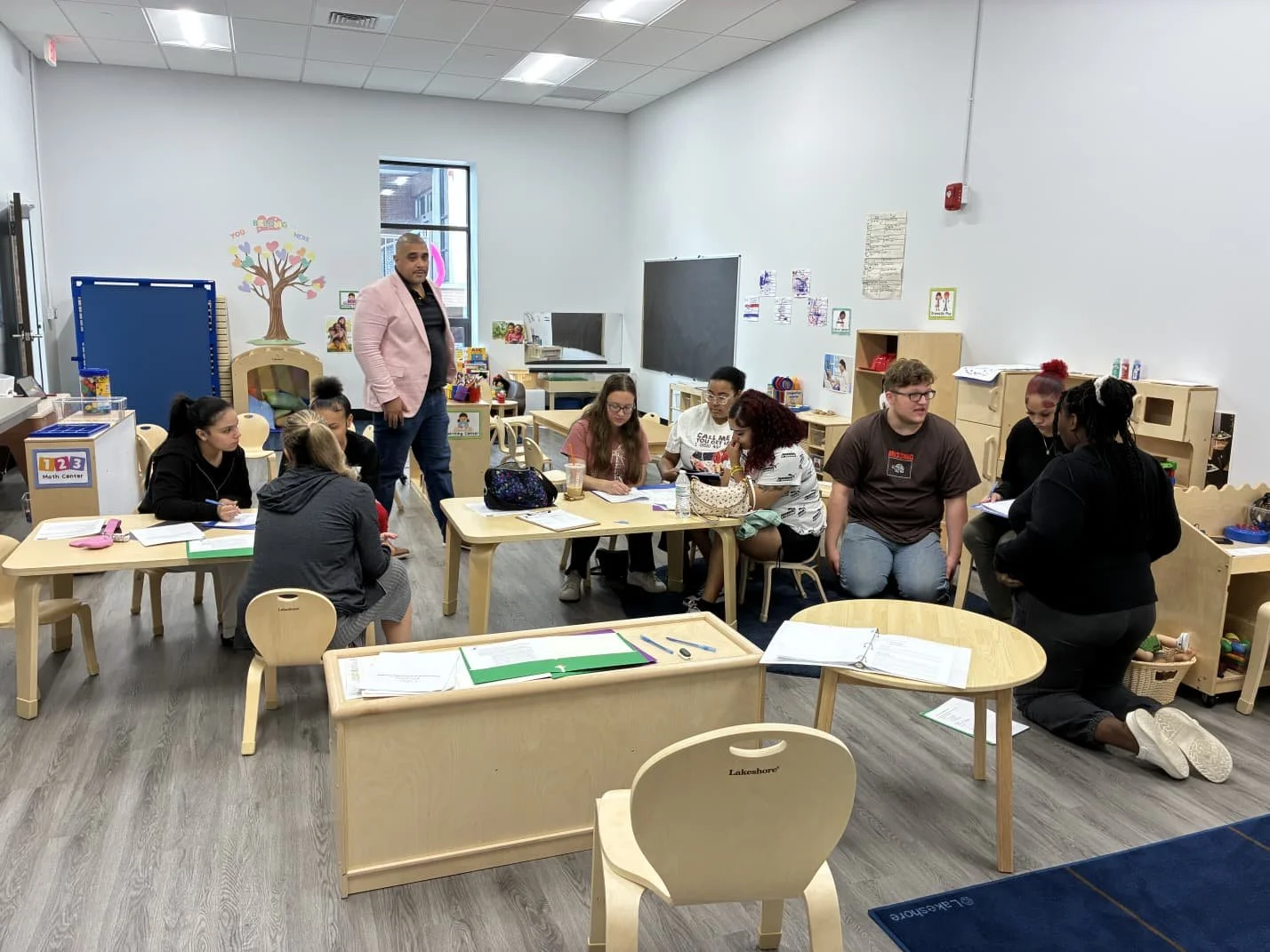 A classroom with several students and an instructor engaged in a group activity. The room has light-colored wooden furniture, educational posters on the walls, and shelves with toys and learning materials.