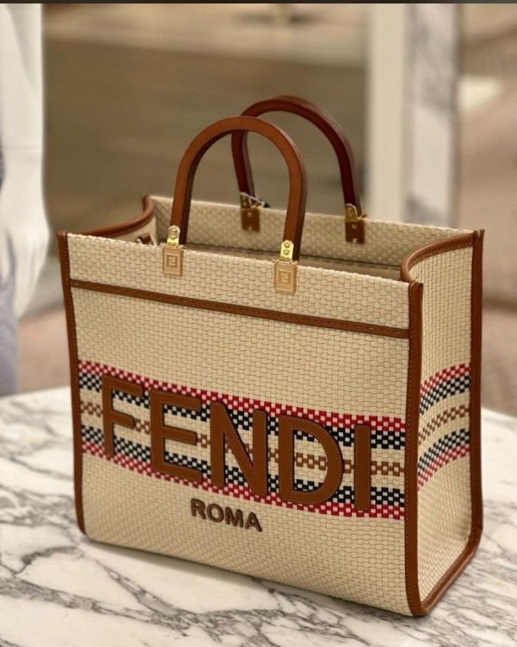 A beige Fendi Roma tote bag with brown leather handles and a red, black, and beige woven design, placed on a marble surface.