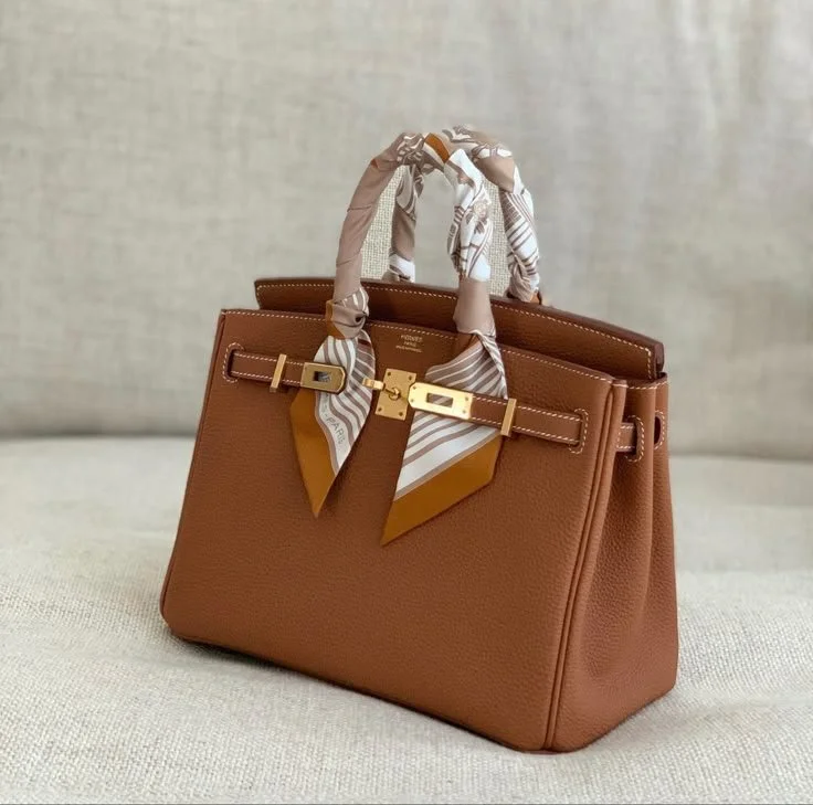 A brown leather handbag with gold hardware and a silk scarf tied around the handle, placed on a neutral surface.