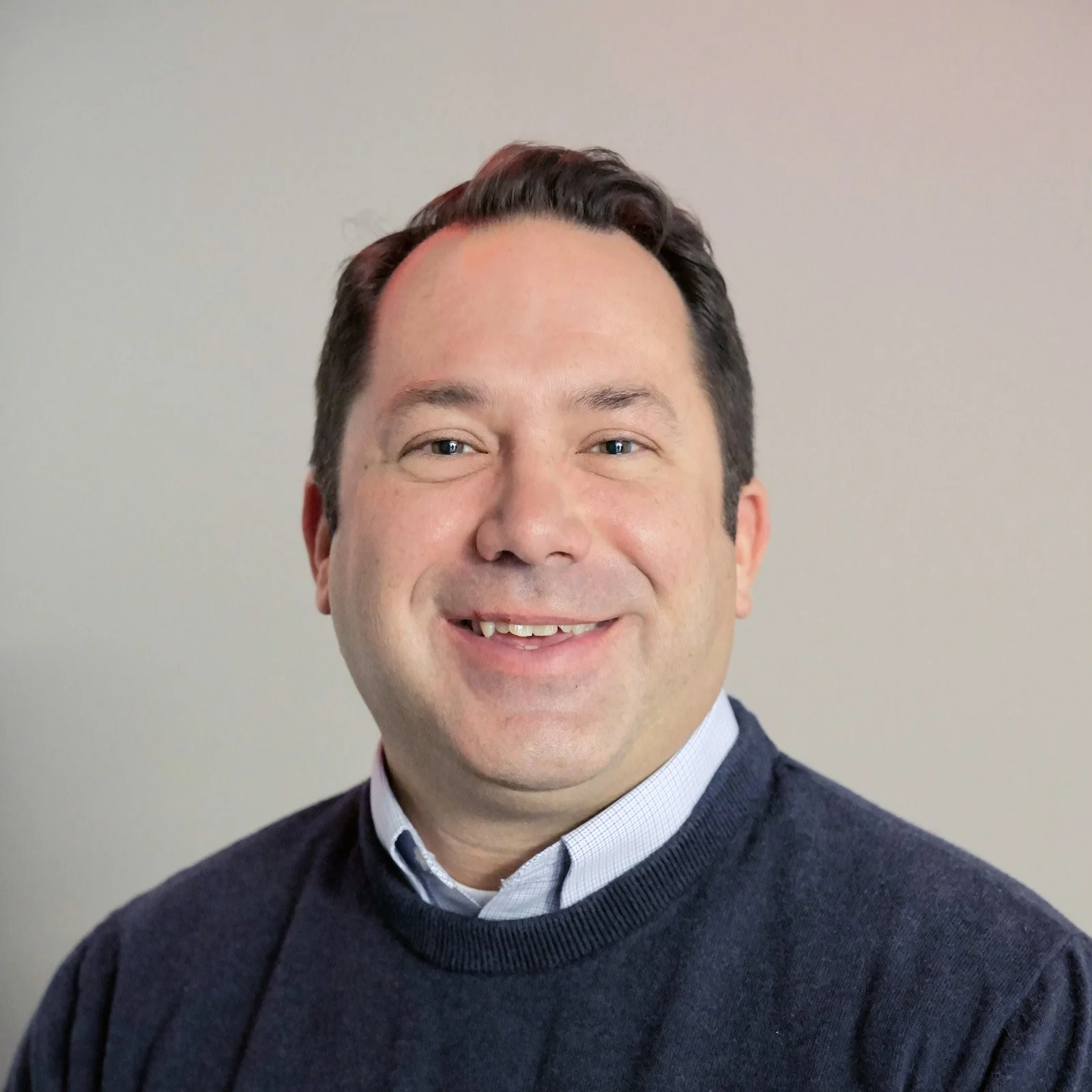 Smiling man with short dark hair wearing a collared shirt and navy sweater, posing against a plain light background.