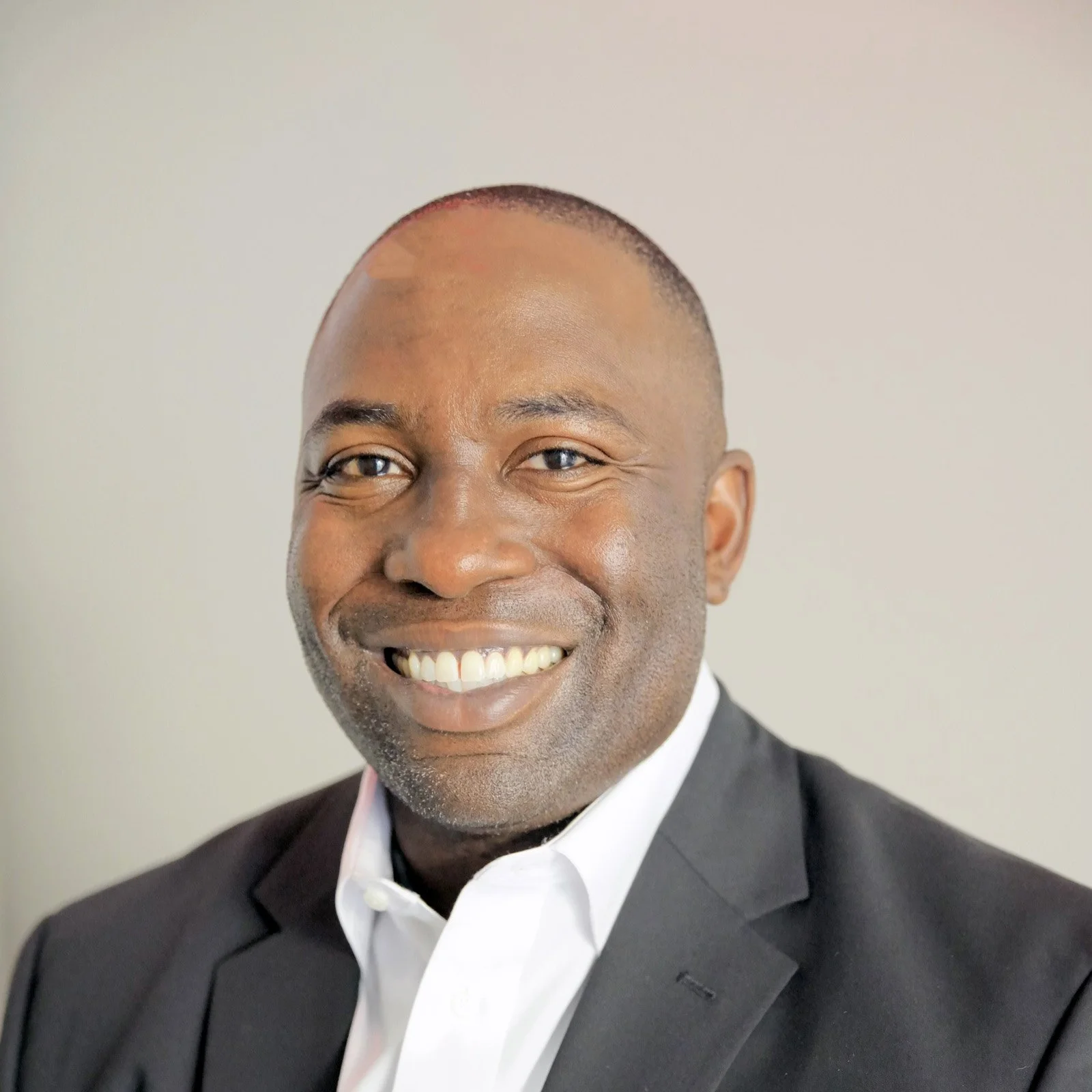 Portrait of a smiling man wearing a white shirt and black suit against a plain background.