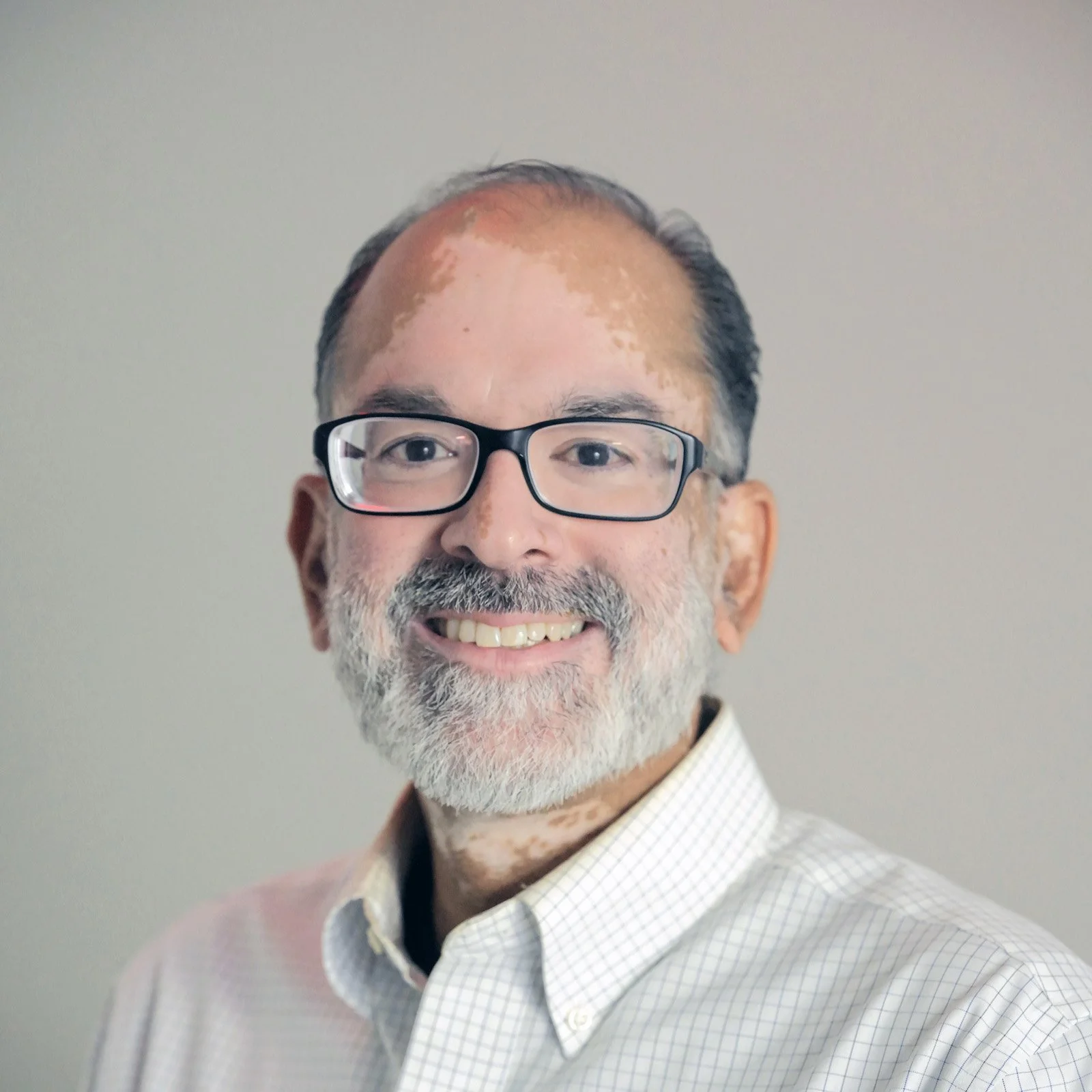A man with glasses, gray beard and short hair wearing a white checkered shirt smiling at the camera.