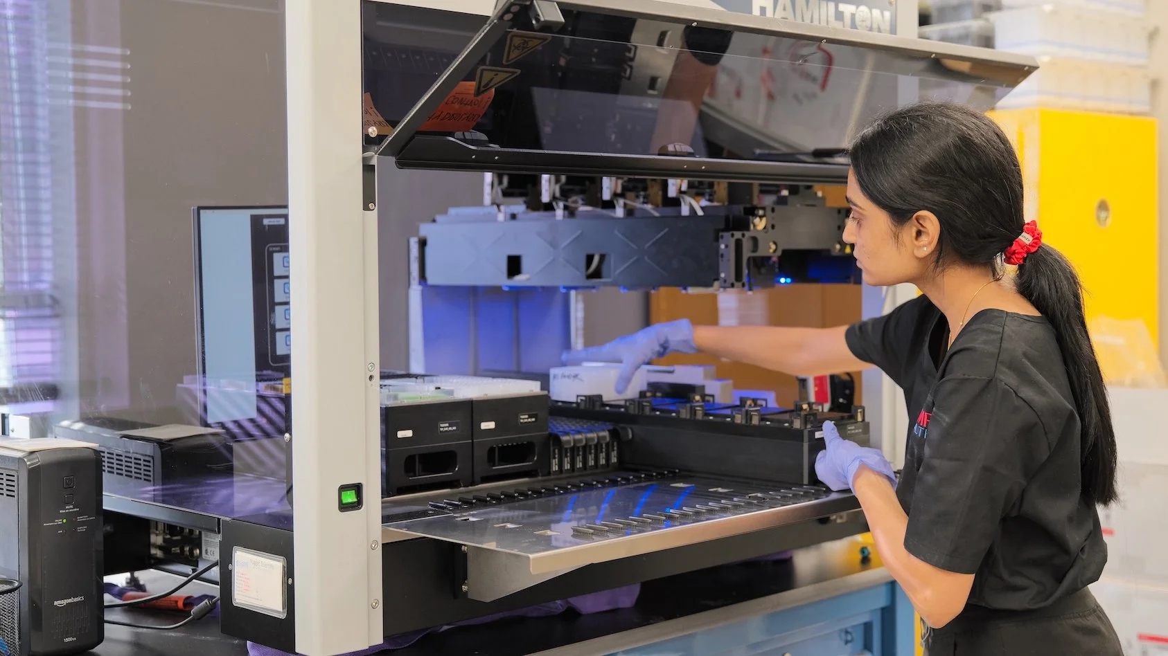 A female scientist working with a scientific instrument in a laboratory. She is wearing gloves and a black uniform, and appears to be handling samples or equipment inside the device.