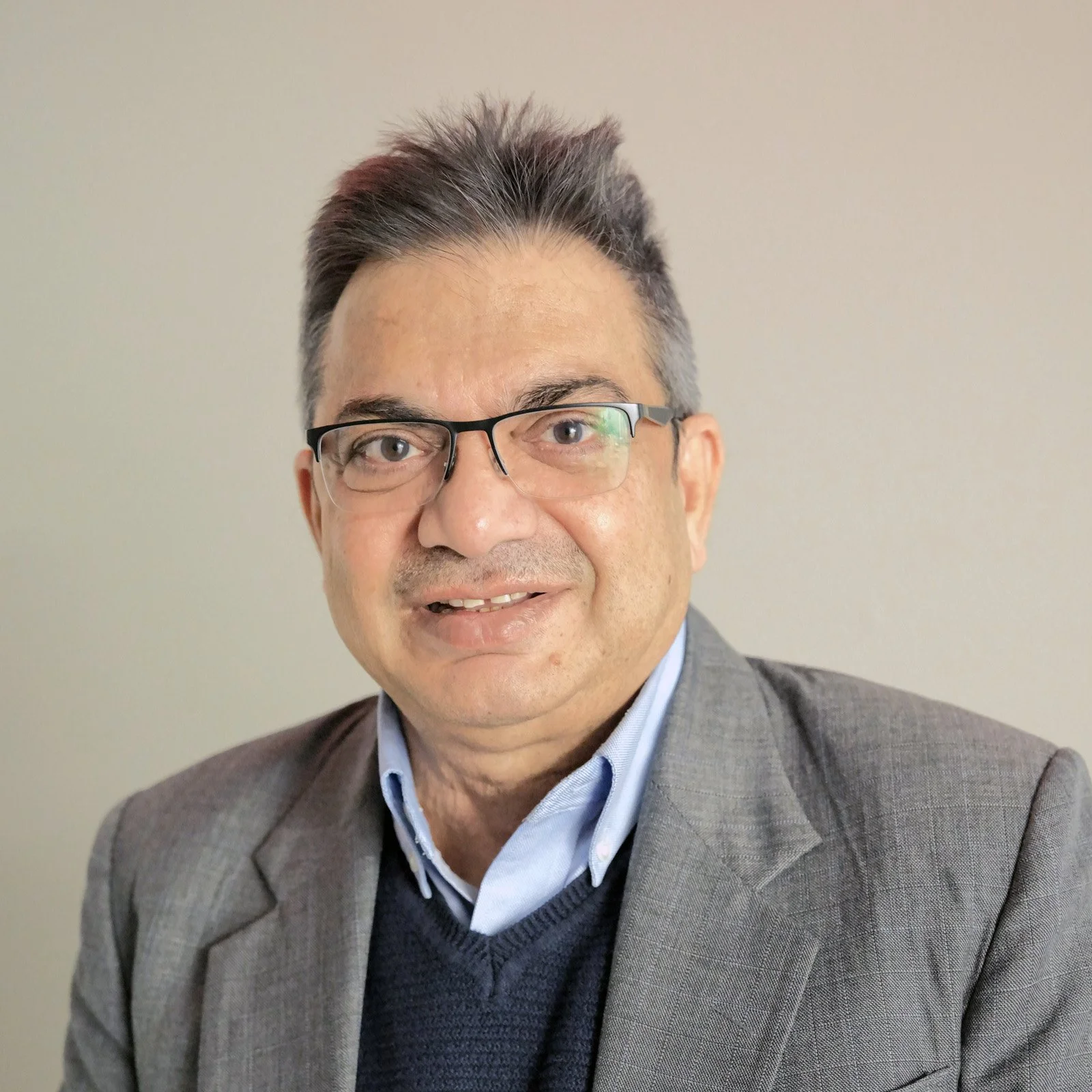 A middle-aged man with glasses, wearing a gray suit jacket, light blue shirt, and navy sweater, smiling against a plain light-colored background.