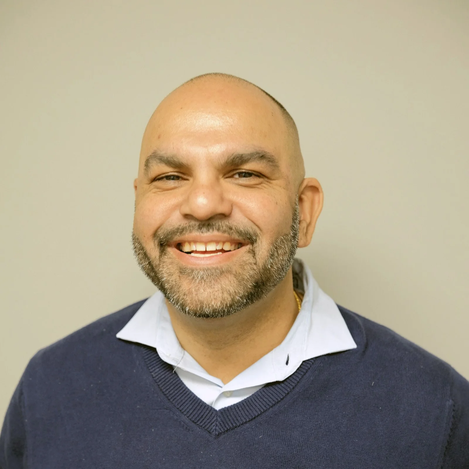 A smiling middle-aged man with a bald head and a beard, wearing a white collared shirt and a dark blue sweater, standing against a neutral background.