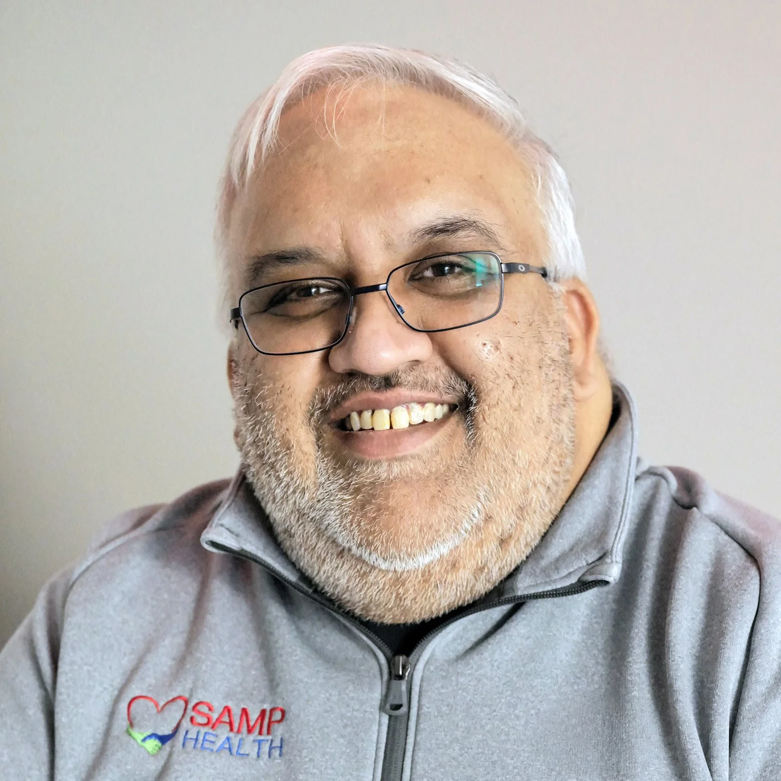 A smiling man with glasses and a gray beard wearing a gray jacket with a SAMP Health logo.