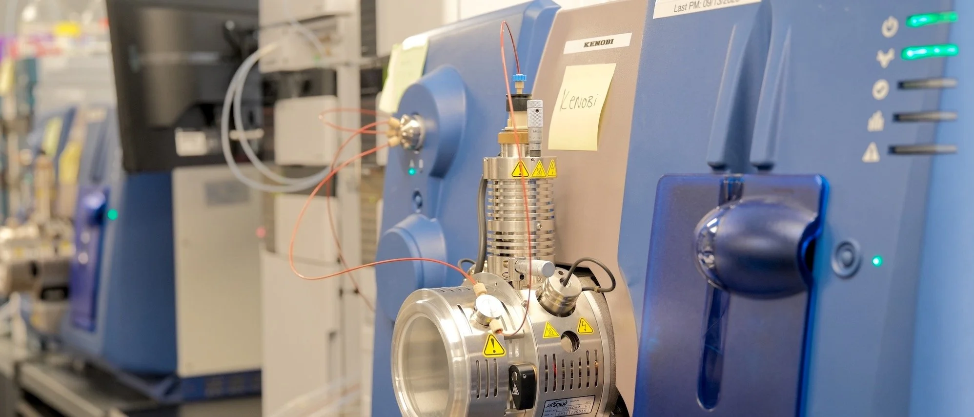 Close-up of a scientific instrument with a blue casing, metal components, warning labels, and connected wires in a laboratory setting.