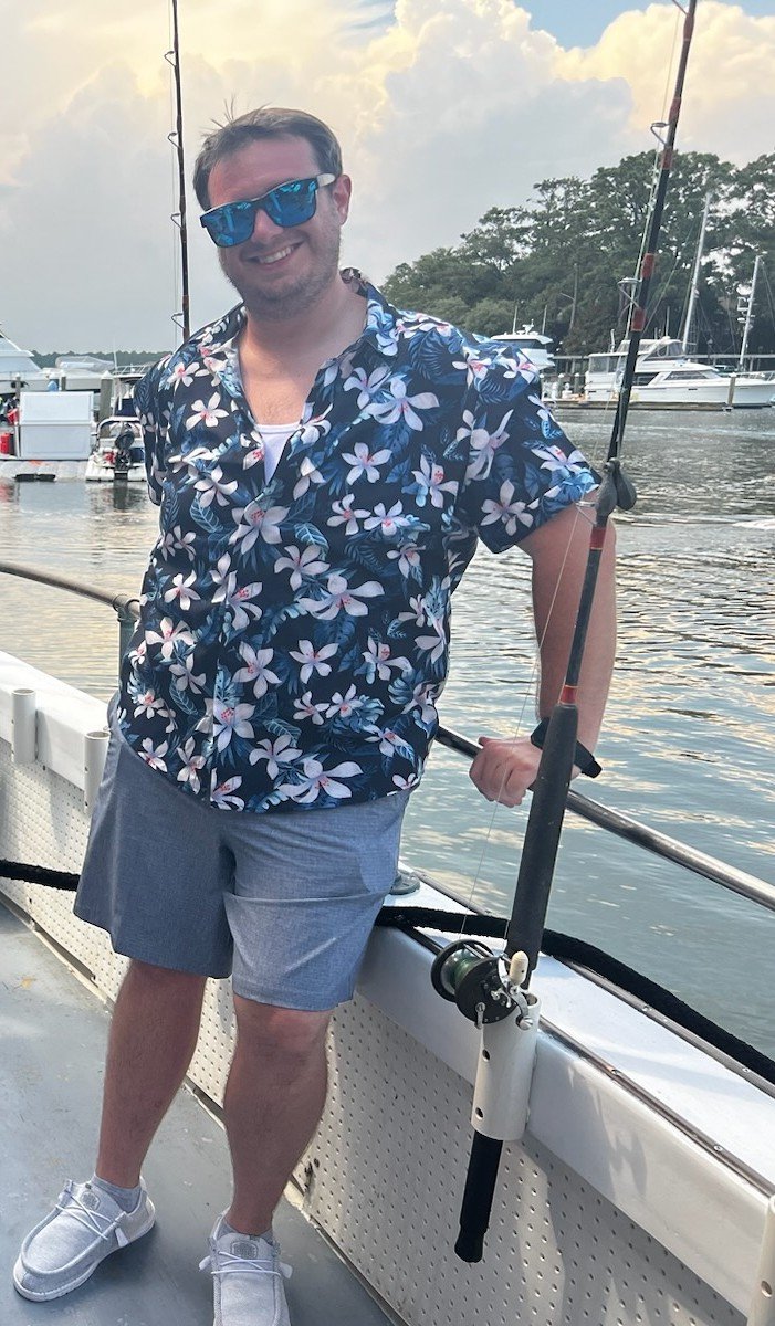 Man in floral shirt and sunglasses standing on a boat with fishing rod by a marina with boats.