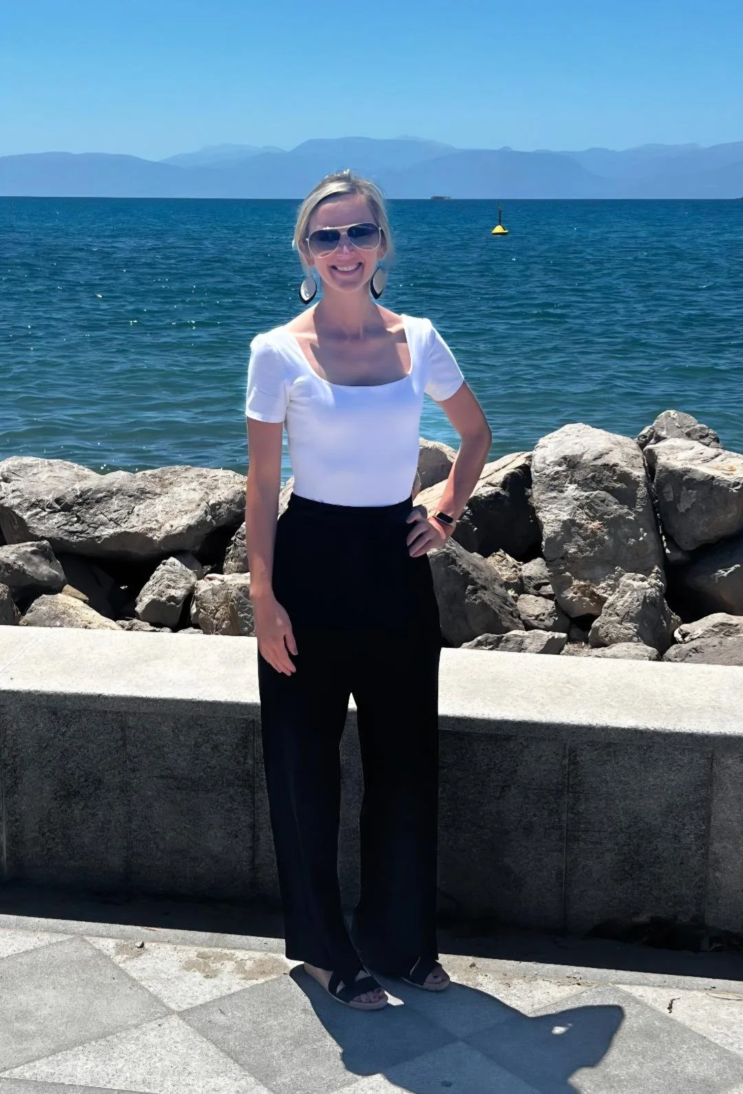 Woman standing on a waterfront promenade beside rocks with an ocean and distant mountains in the background, wearing sunglasses, a white top, and black pants.
