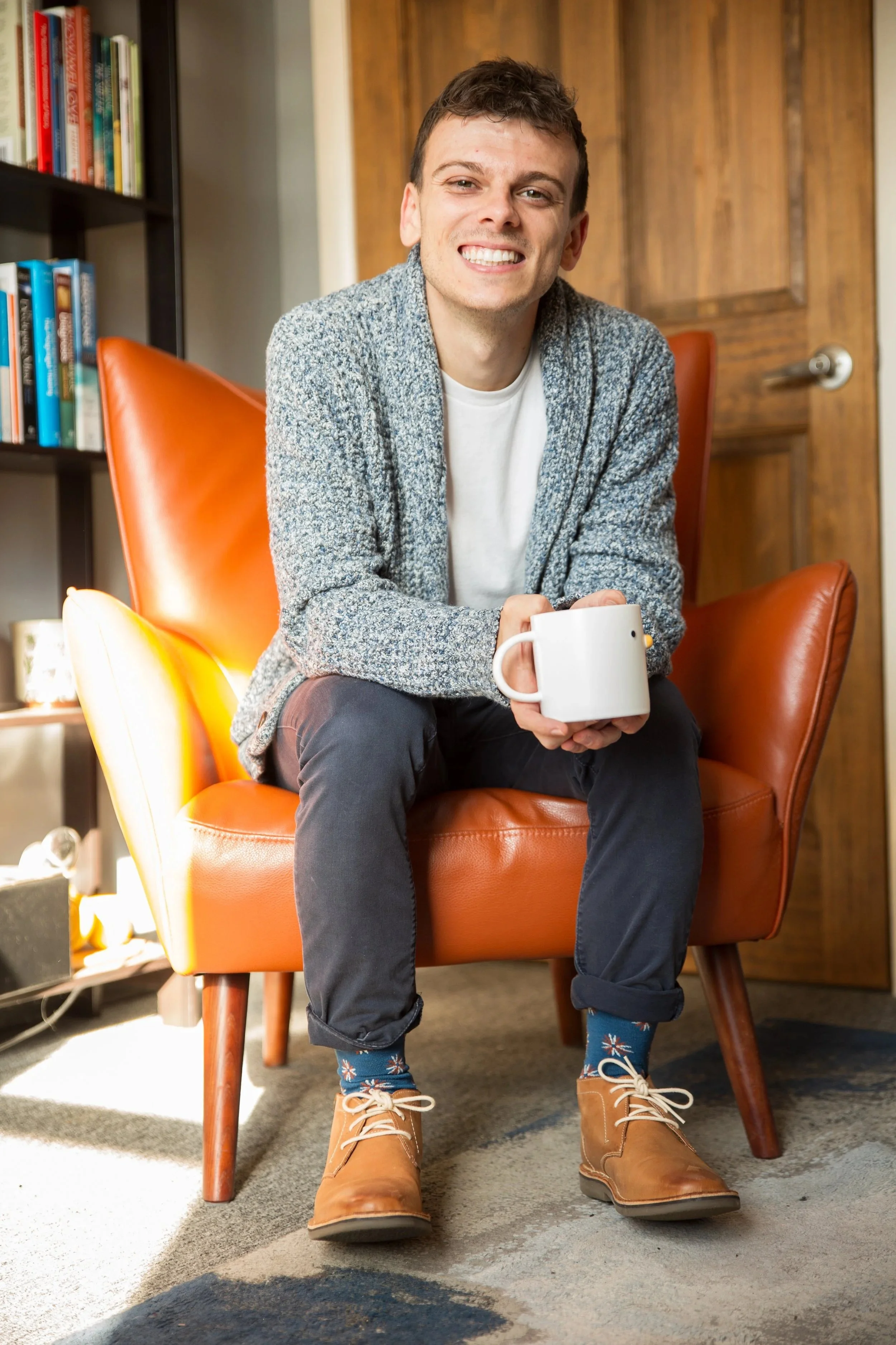 A young man with a big smile sitting on an orange armchair, holding a white mug. He is wearing a gray sweater, dark pants, tan boots, and colorful socks. Behind him are bookshelves and a wooden door.