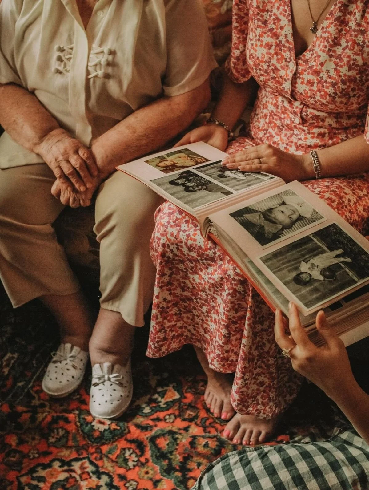 Two women are sitting on a colorful patterned rug, looking at a photo album together. One woman is elderly, wearing a light-colored blouse and beige pants, with white sneakers. The other woman, younger, is wearing a red and white floral dress and has bare feet. They are examining black-and-white and color photographs in the album.