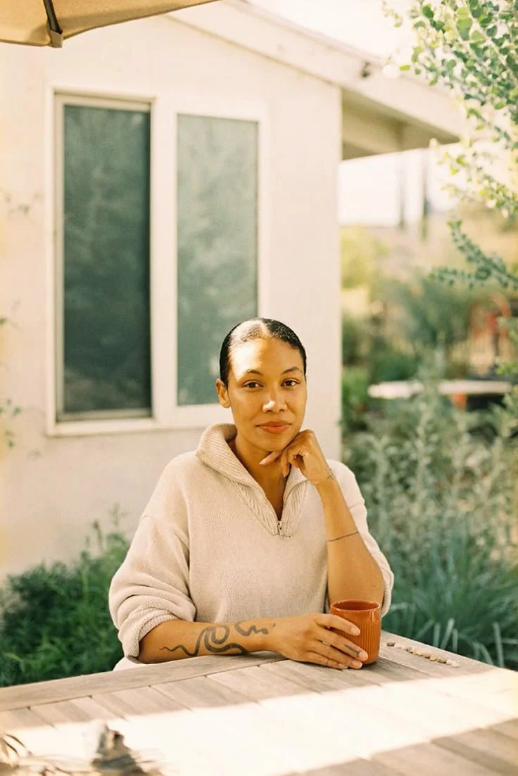 Woman sitting at a table outside, holding a coffee mug, with a house and greenery in the background.