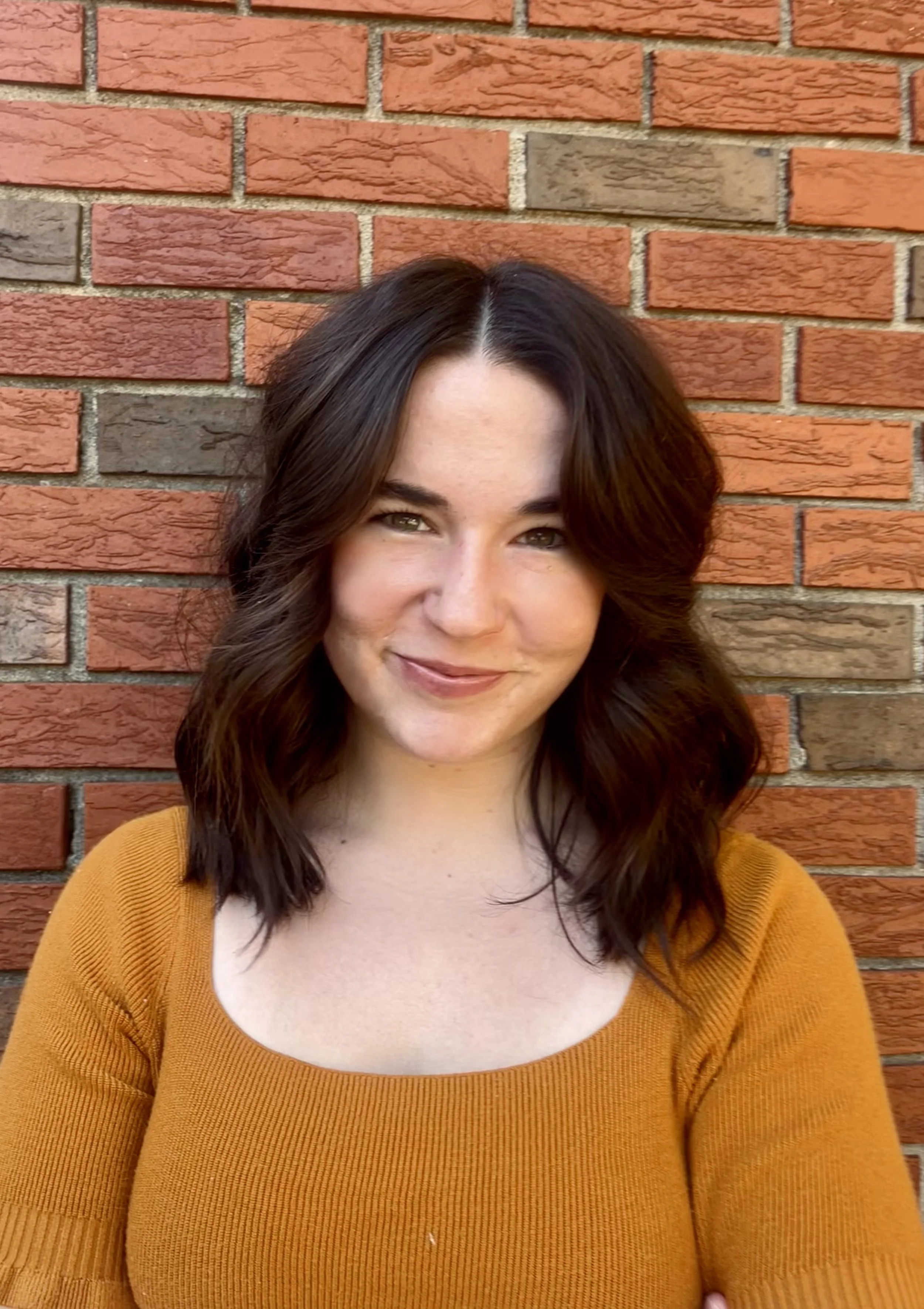 A young woman with shoulder-length dark brown hair smiling in front of a red brick wall, wearing a mustard yellow top.