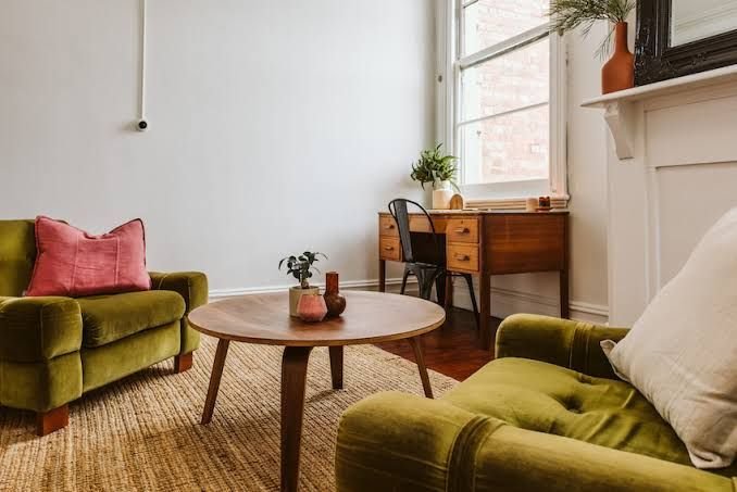 Therapy office with green velvet sofas, a round wooden coffee table, a wooden desk with a black chair, a window with white blinds, a potted plant, and decorative vases.