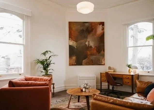 Therapy office with white walls, two large windows, a brown leather sofa, a matching armchair, a wooden coffee table with flowers, a wooden desk with a brown chair, a large abstract painting, a potted plant, and a ceiling light