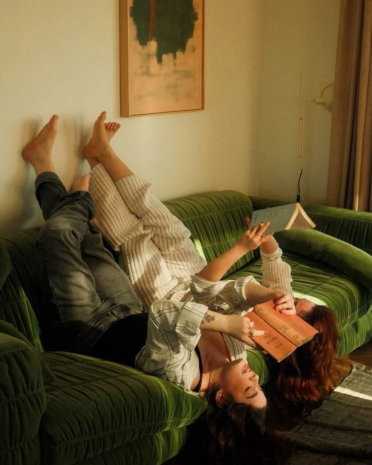 Two women are lying upside down on a green sofa, reading books in a cozy living room with a framed picture on the wall.