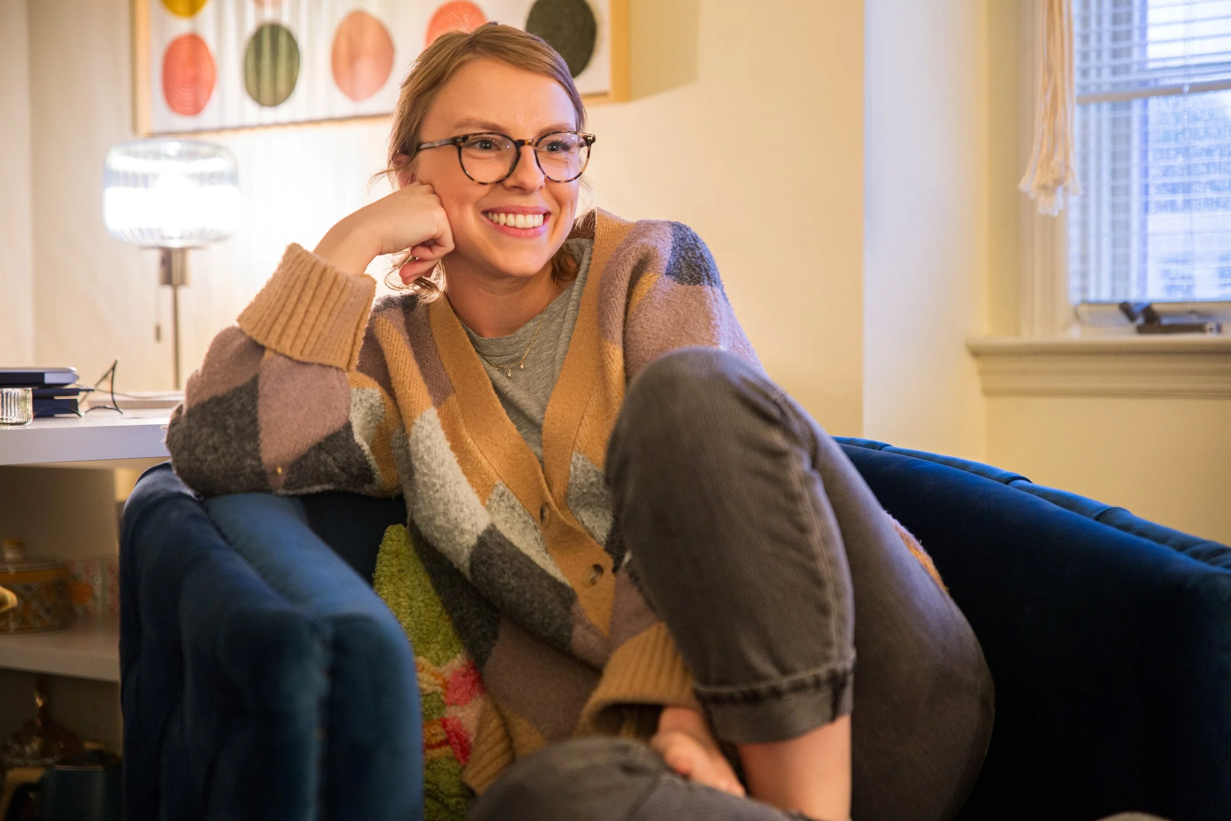 A woman with glasses smiling while sitting on a dark blue couch in a cozy living room, leaning her head on her hand with her legs crossed. She is wearing a multicolored, patterned cardigan and grey pants. There is a white wall with a framed art piece and a window with blinds in the background.