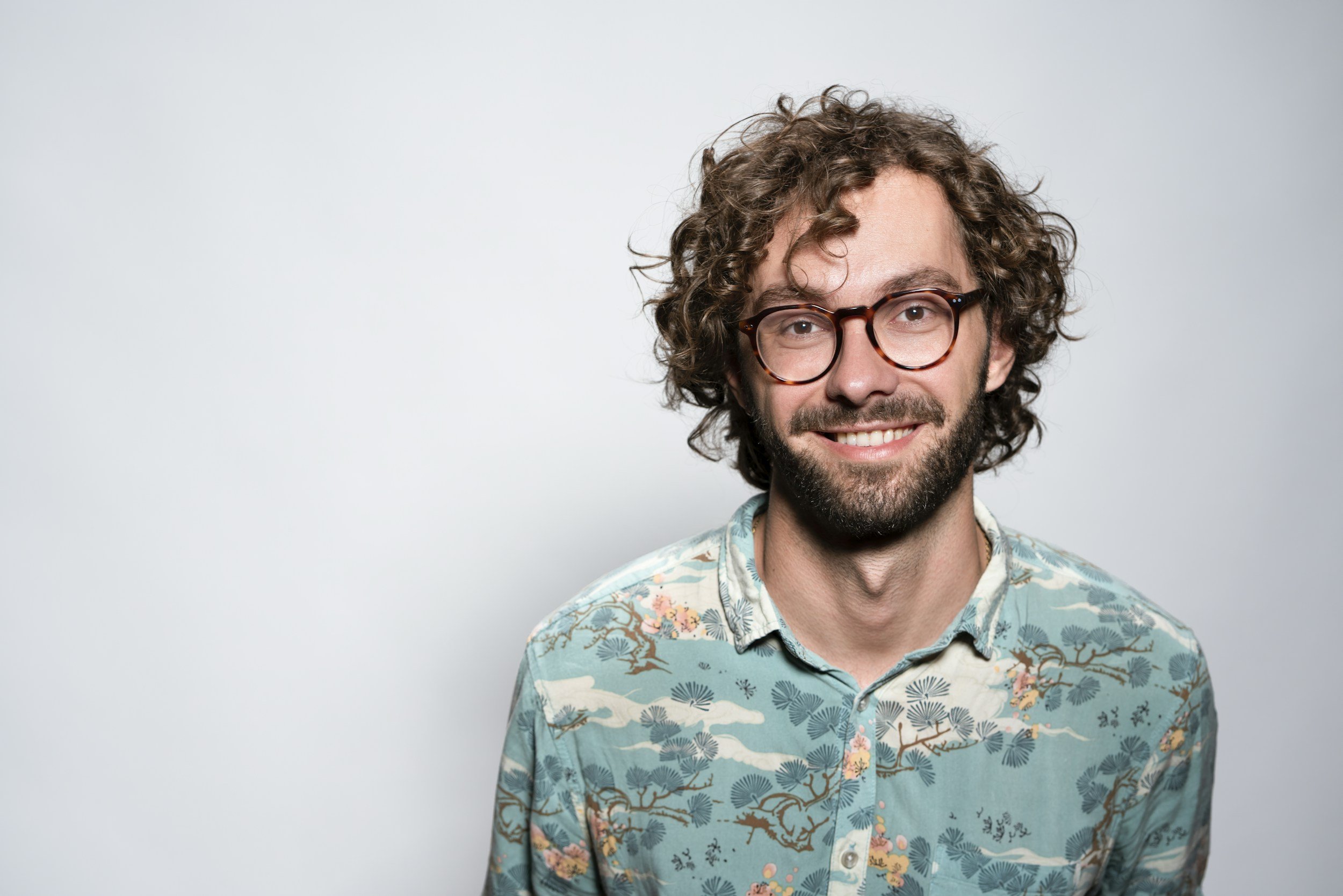 A smiling man with curly hair, glasses, a beard, and a floral patterned shirt standing against a plain light gray background.