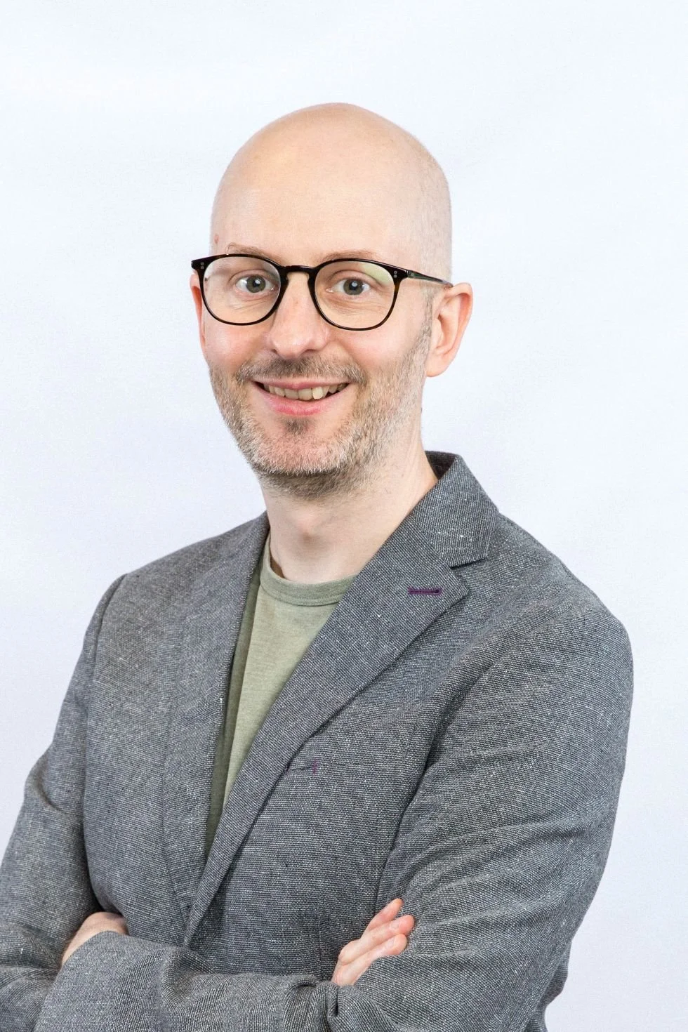 A smiling man with glasses, a light beard, wearing a gray blazer over a beige shirt with a light background.
