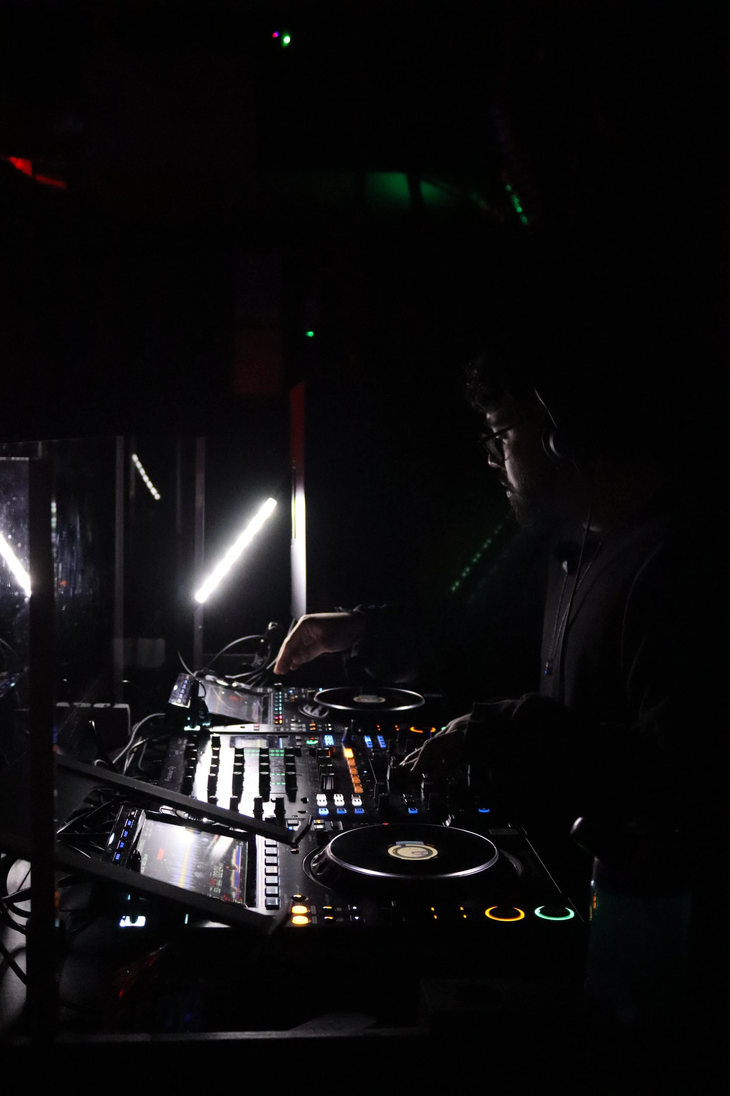 A DJ is performing with equipment, wearing headphones, in a dark environment illuminated by the colorful lights on the DJ gear.