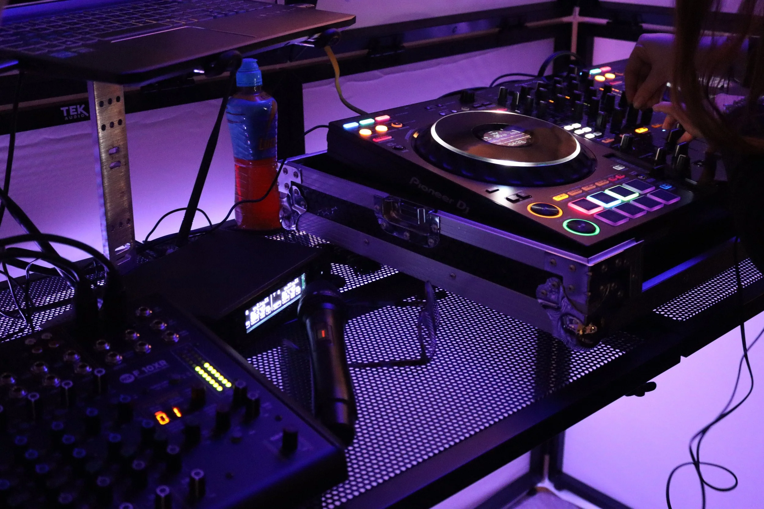 A DJ setup with Pioneer DJ equipment including a mixer and a laptop, with hands adjusting controls, in a dimly lit environment.