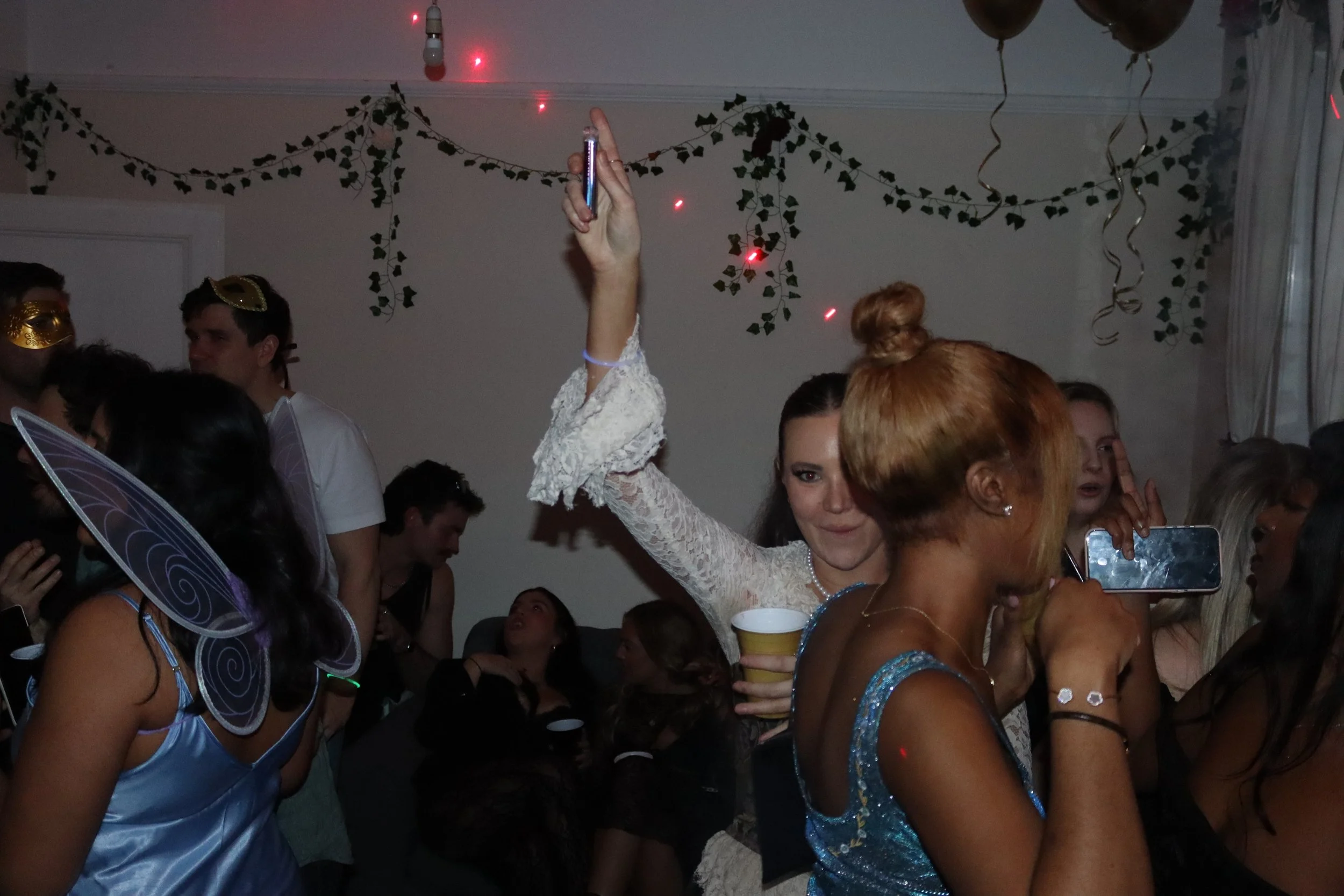People at a party wearing costumes, some with fairy wings and mask accessories, crowded together, holding drinks, with hanging decorations and balloons in the background.