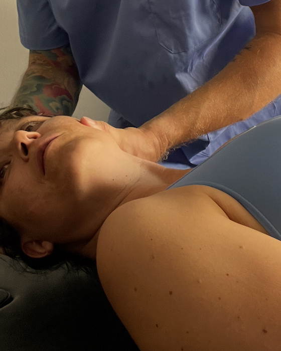 Paciente acostada en una camilla recibiendo una consulta médica, con un profesional de la salud revisando su cuello.